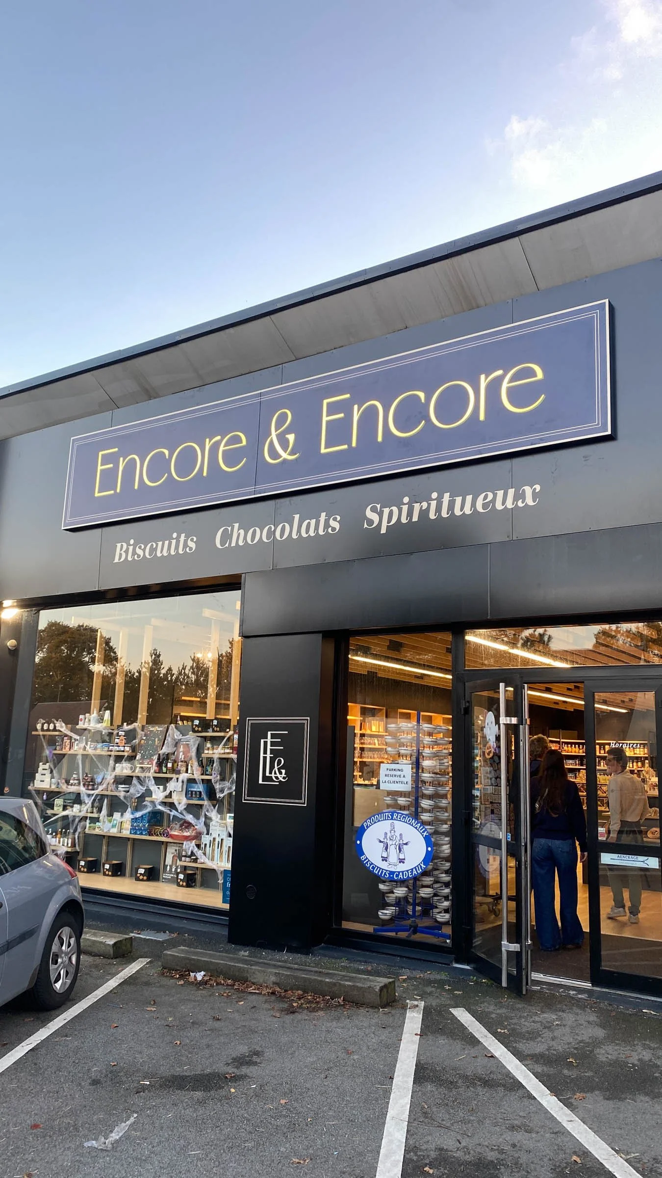 Façade d'une boutique nommée 'Encore & Encore', vendant des biscuits, chocolats et produits spiritueux, avec des clients à l'intérieur et des voitures dans le parking extérieur.