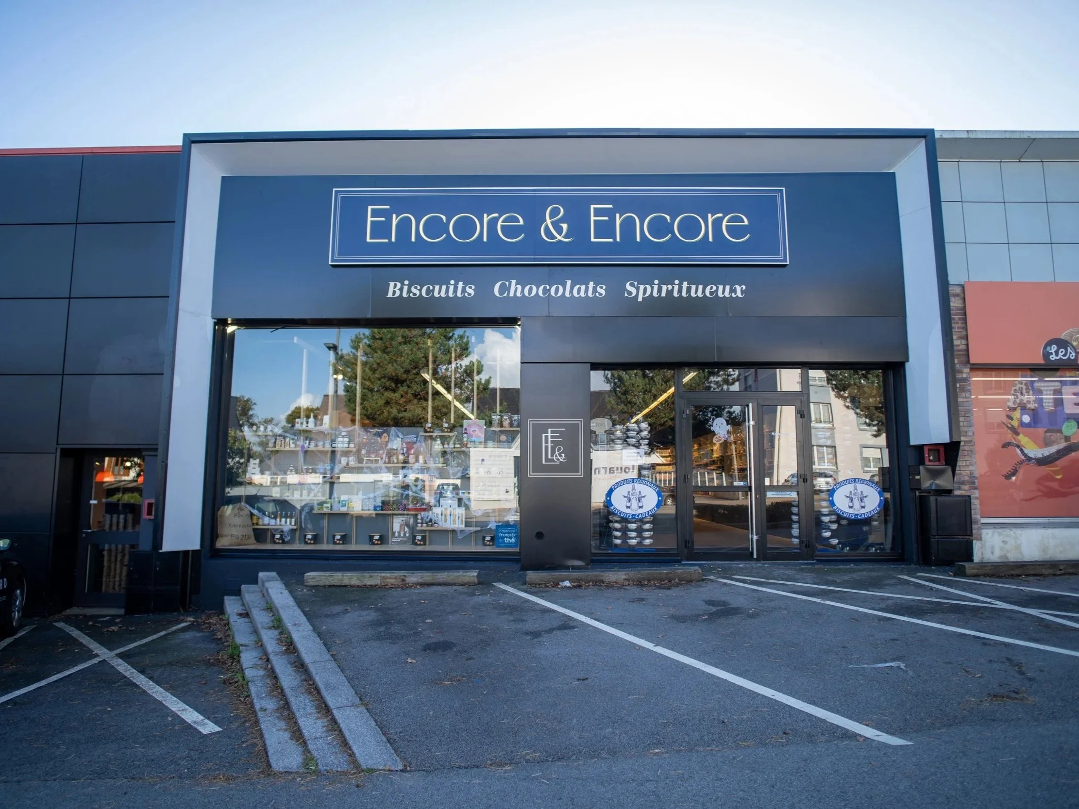 Fenêtre du magasin Encore & Encore avec enseigne, proposant biscuits, chocolats, et spiritueux, dans un centre commercial avec parking vide en avant, jour ensoleillé.