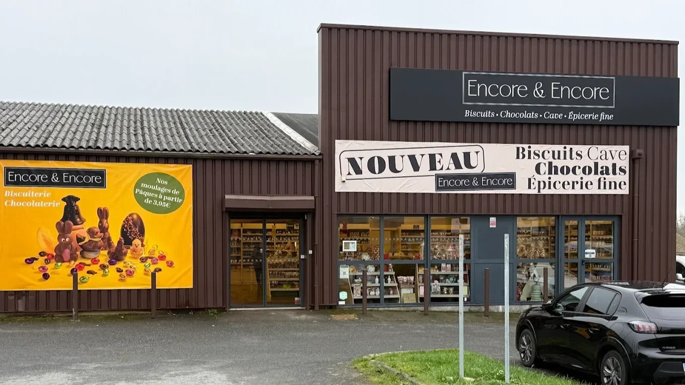 Une boutique nommée 'Encore & Encore' vendant biscuits, chocolats, cave et épicerie fine. La devanture comporte une grande bannière annonçant un nouveau produit ou service. La façade est en bois brun avec de grandes vitres. Un parking est visible avec une voiture noire garée devant.