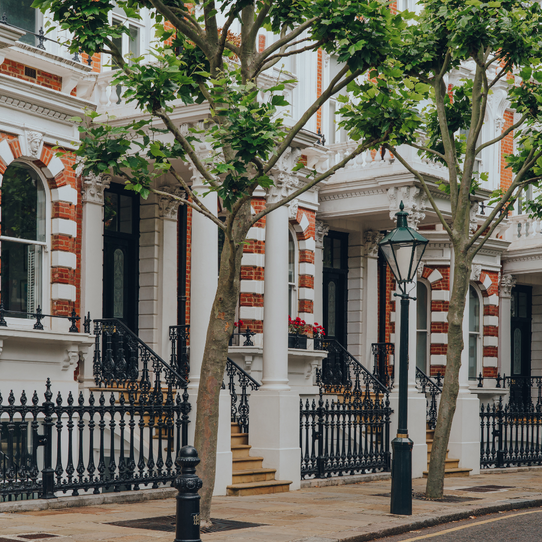 A row of elegant townhouses with ornate architecture, white columns, red brick accents, black iron railings, stairs leading to entrances, trees lining the sidewalk, and a street lamp.