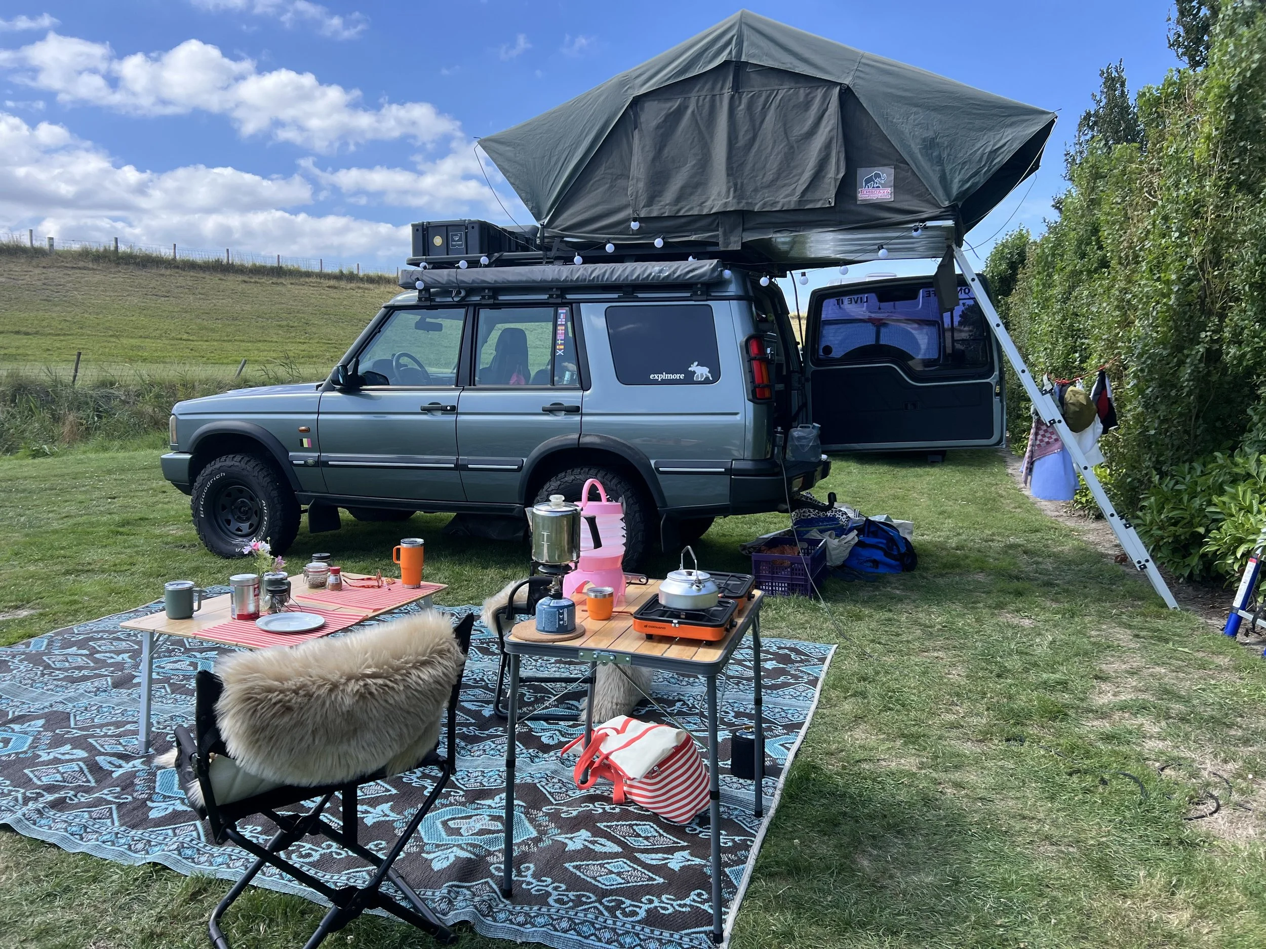 Camperpark met een omgebouwde auto met uitklapbare tent, buiten een gespreid tapijt met kampeerbenodigdheden, stoelen en een tafel in een groen veld onder een blauwe lucht met wolken.