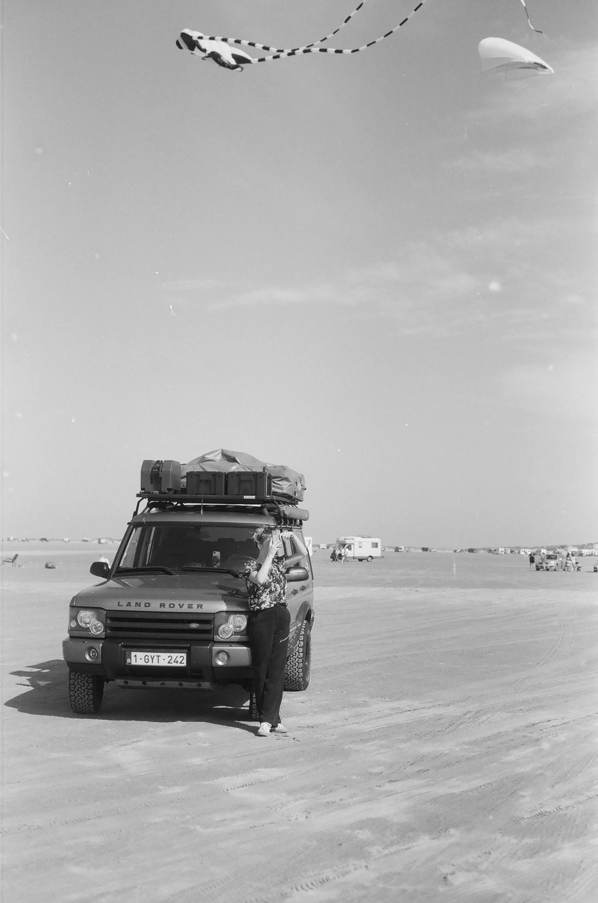 Een woman staat naast een Land Rover op een uitgestrekte, zandachtige vlakke ondergrond, met een caravan op de achtergrond. In de lucht vliegen twee gekleurde vliegers.