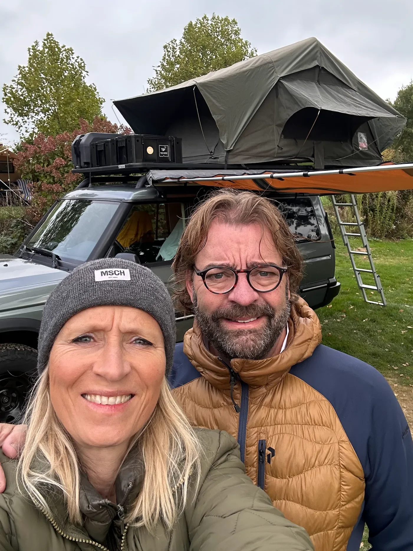 Twee mensen maken een selfie voor een camper met een daktent, op een grasveld met bomen op de achtergrond.