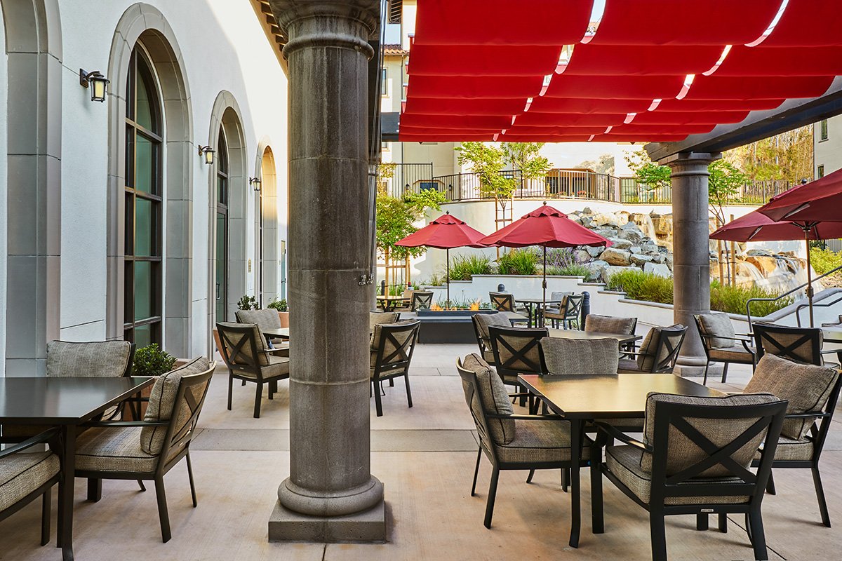 An shaded outdoor lounge with fire table sits between the pool building and a serene, cascading water feature.