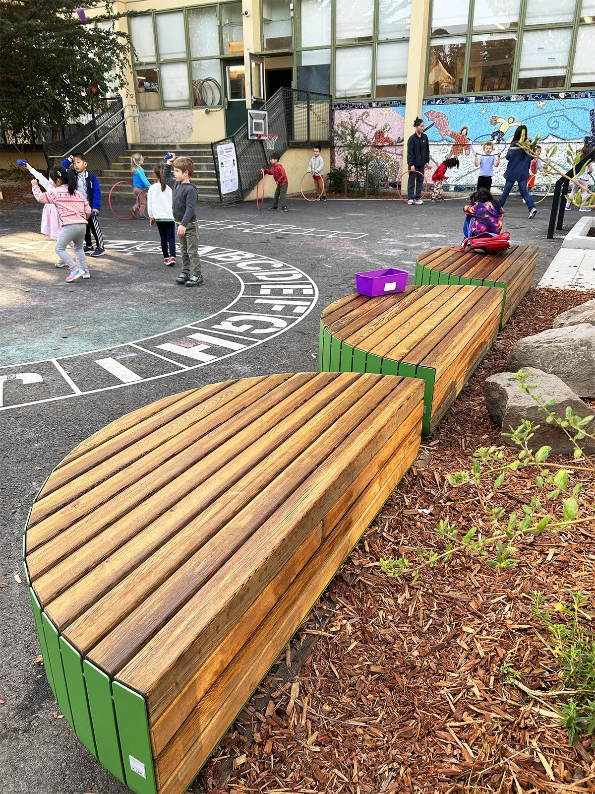 Curved wood benches introduce warmth, texture, and flexible seating opportunities along the play yard edge, creating informal spaces for gathering, observation, and outdoor engagement.