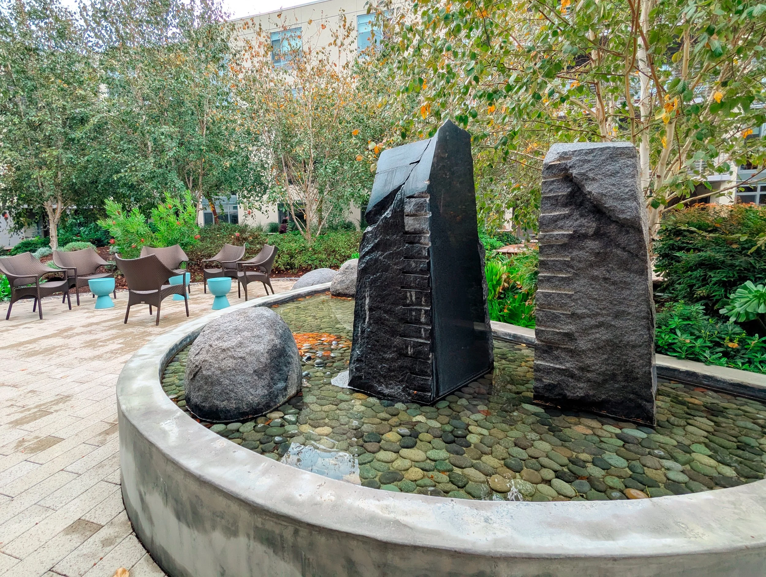 Water glides across stone and rock in the courtyard's central fountain. Framed by shrubs and trees, this focal element offers serene sights and sounds.
