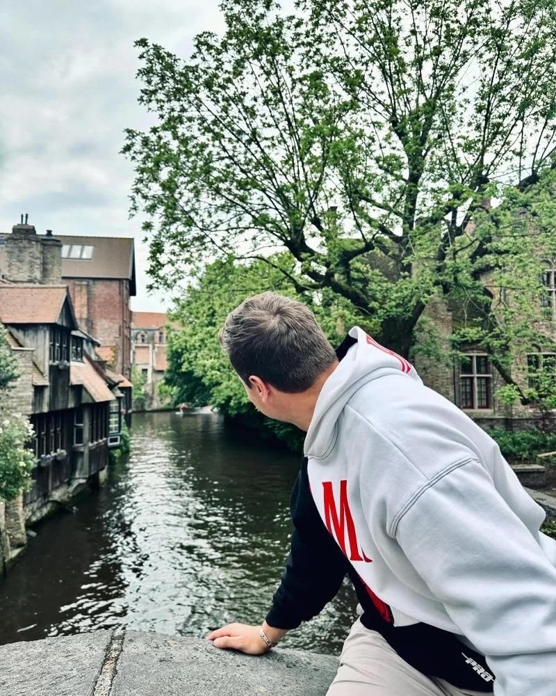 A young man in a white hoodie leaning over a bridge to look at a canal with houseboats, trees, and older buildings in the background on a cloudy day.