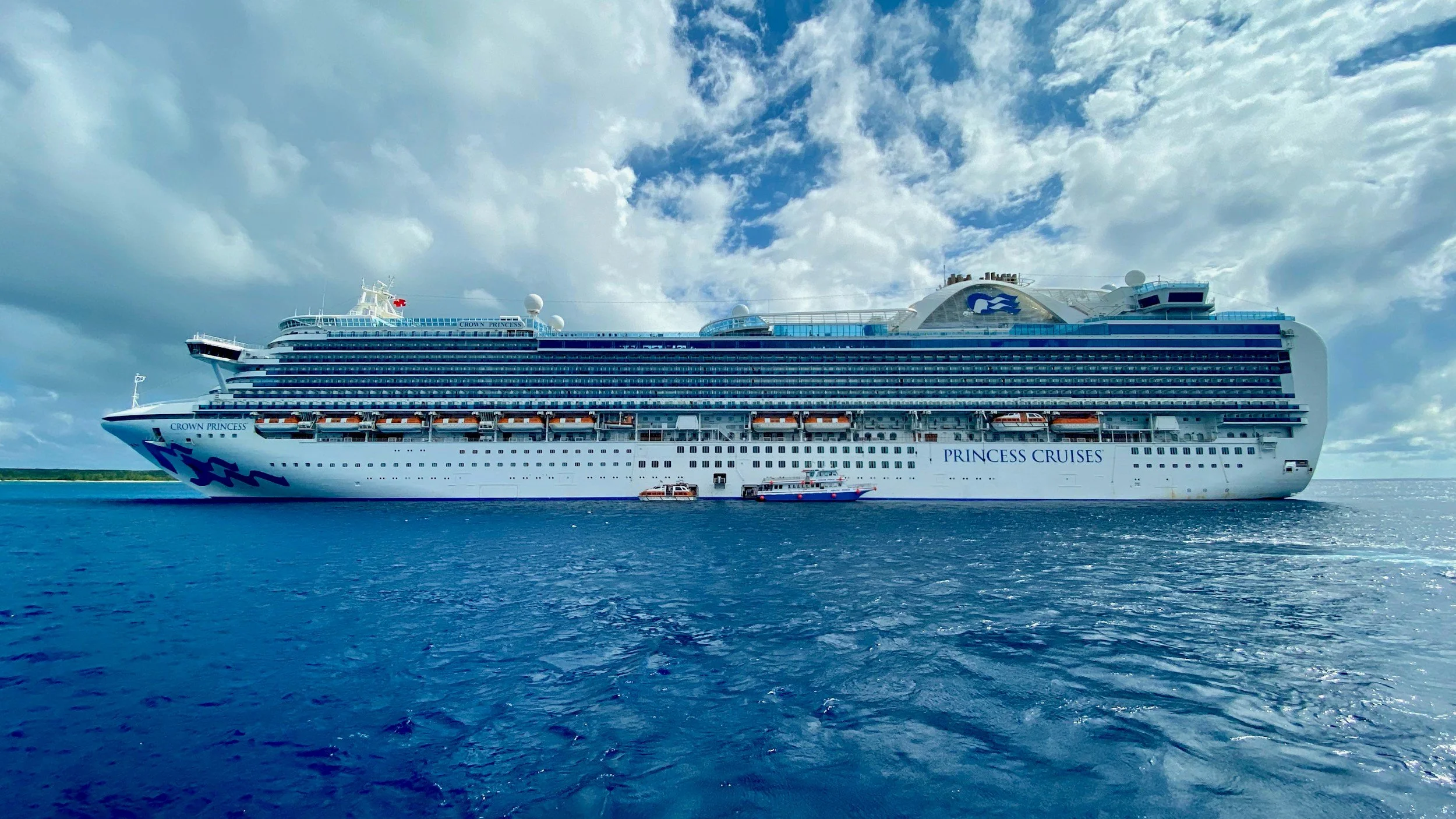 A large white cruise ship labeled Princess Cruises and Crown Princess is sailing in the ocean with a partly cloudy sky above.