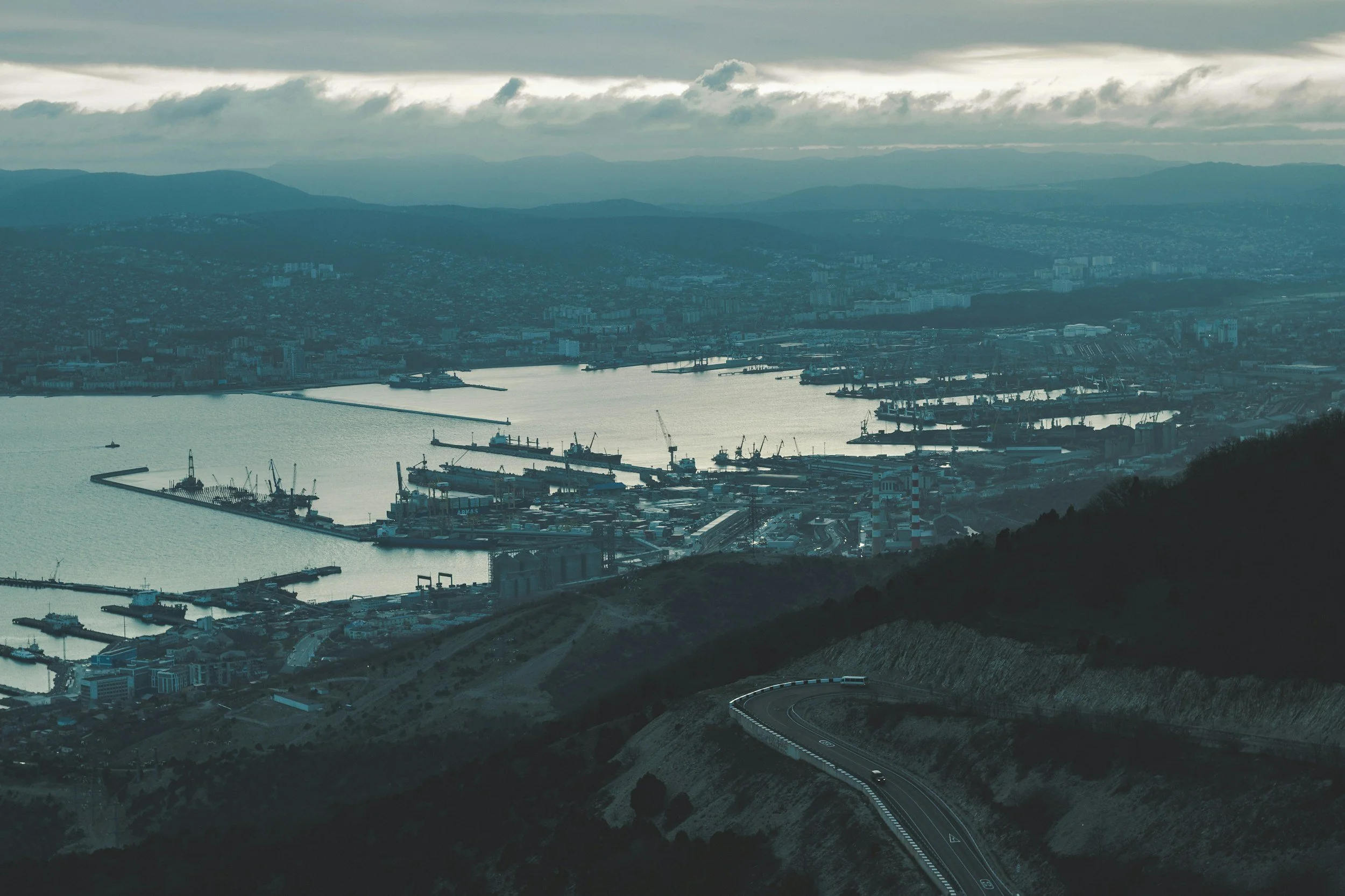 A view of a harbor with ships and cranes from a mountainous area, with a winding road in the foreground and cityscape in the background under a cloudy sky.
