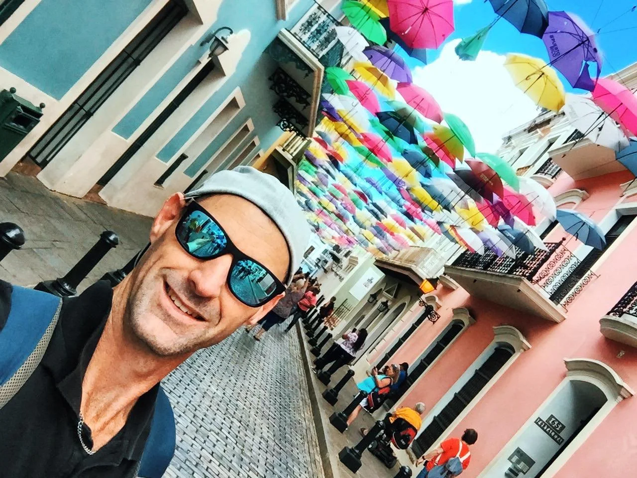 A man wearing sunglasses, a gray cap, and a backpack taking a selfie on a colorful street with numerous umbrellas hanging overhead and other people in the background.