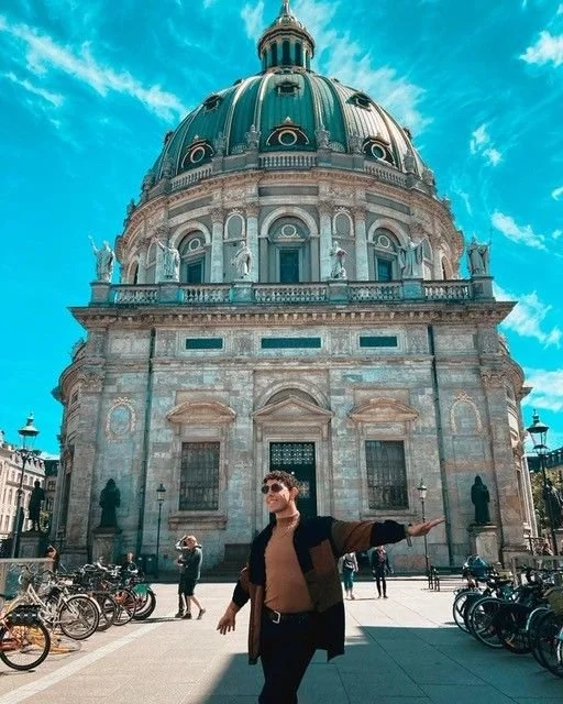 Person in black coat and beige sweater standing in front of large historic building with a green dome and statues, under cloudy blue sky.