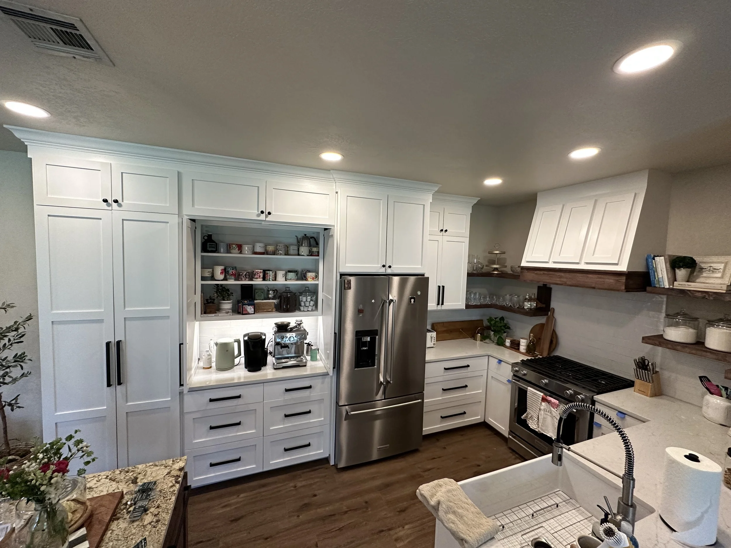 A kitchen with white cabinets, stainless steel refrigerator, stove, and open shelving with cups and jars. Overhead lighting and a granite countertop near a sink with basket and towel.