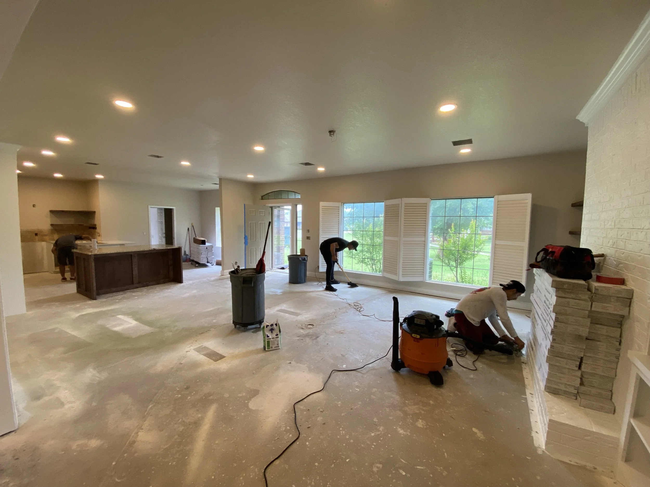 Living room under renovation with workers, construction tools, stacked boxes, and large windows showing a green yard outside.