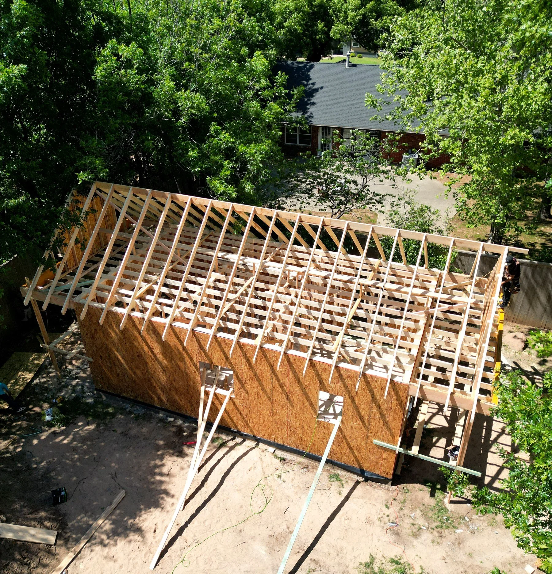 View of a house under construction with a wooden roof framework, surrounded by trees and a yard.