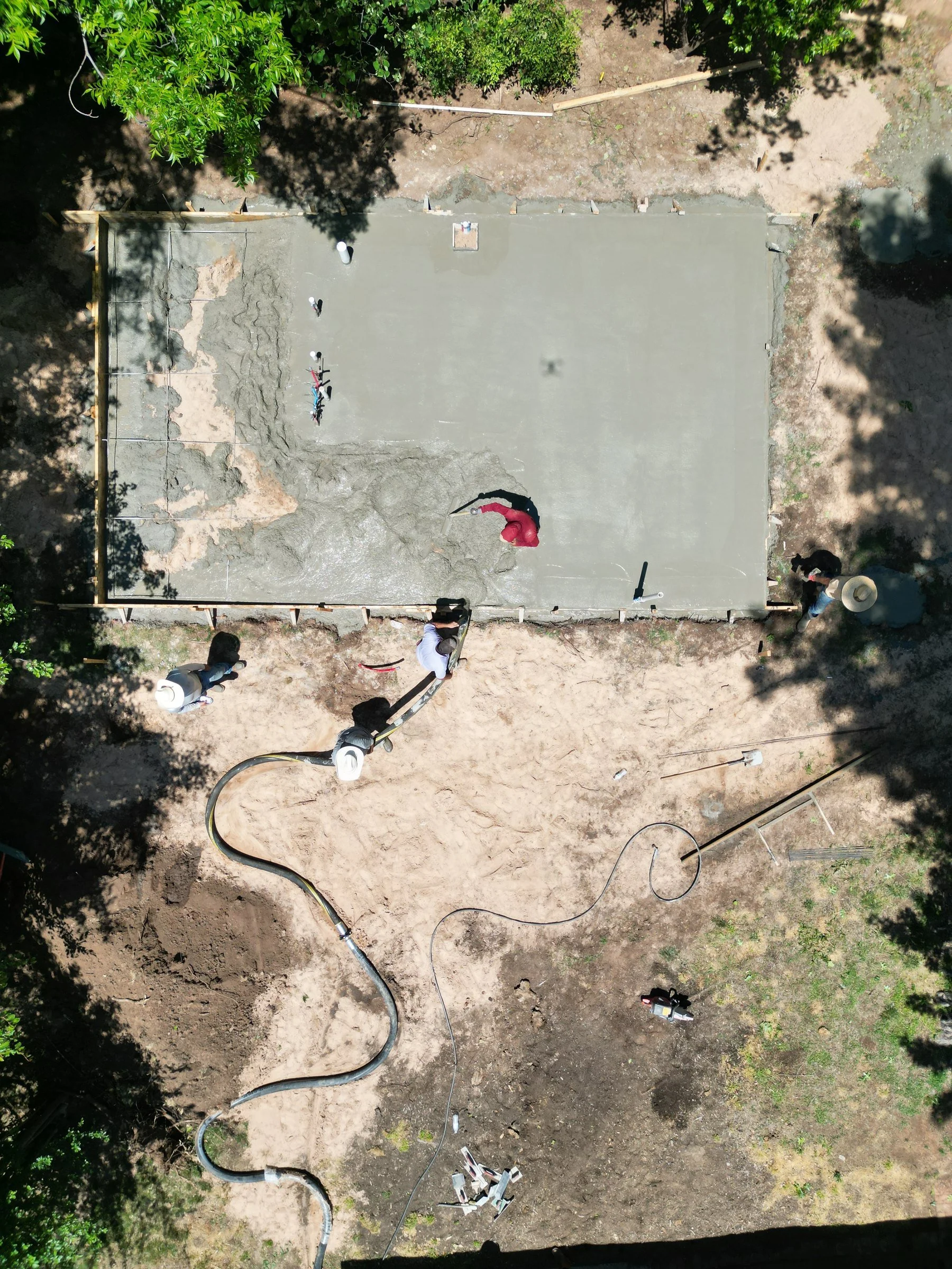 Overhead view of a construction site where workers are pouring concrete for a small slab, with surrounding dirt, trees, and construction tools and hoses nearby.