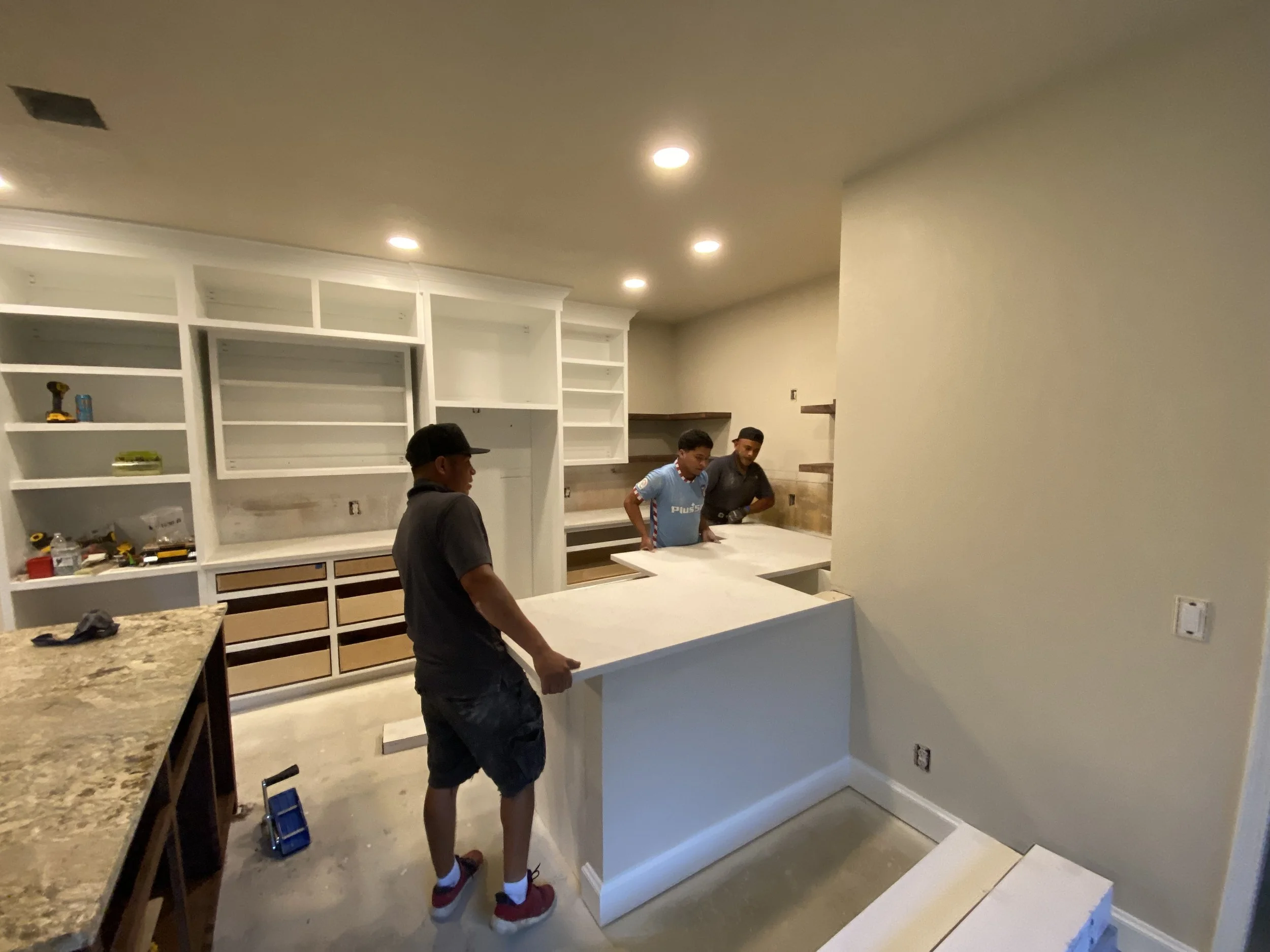 Three men working on a kitchen renovation, installing a kitchen island, with built-in shelves and cabinetry in the background.