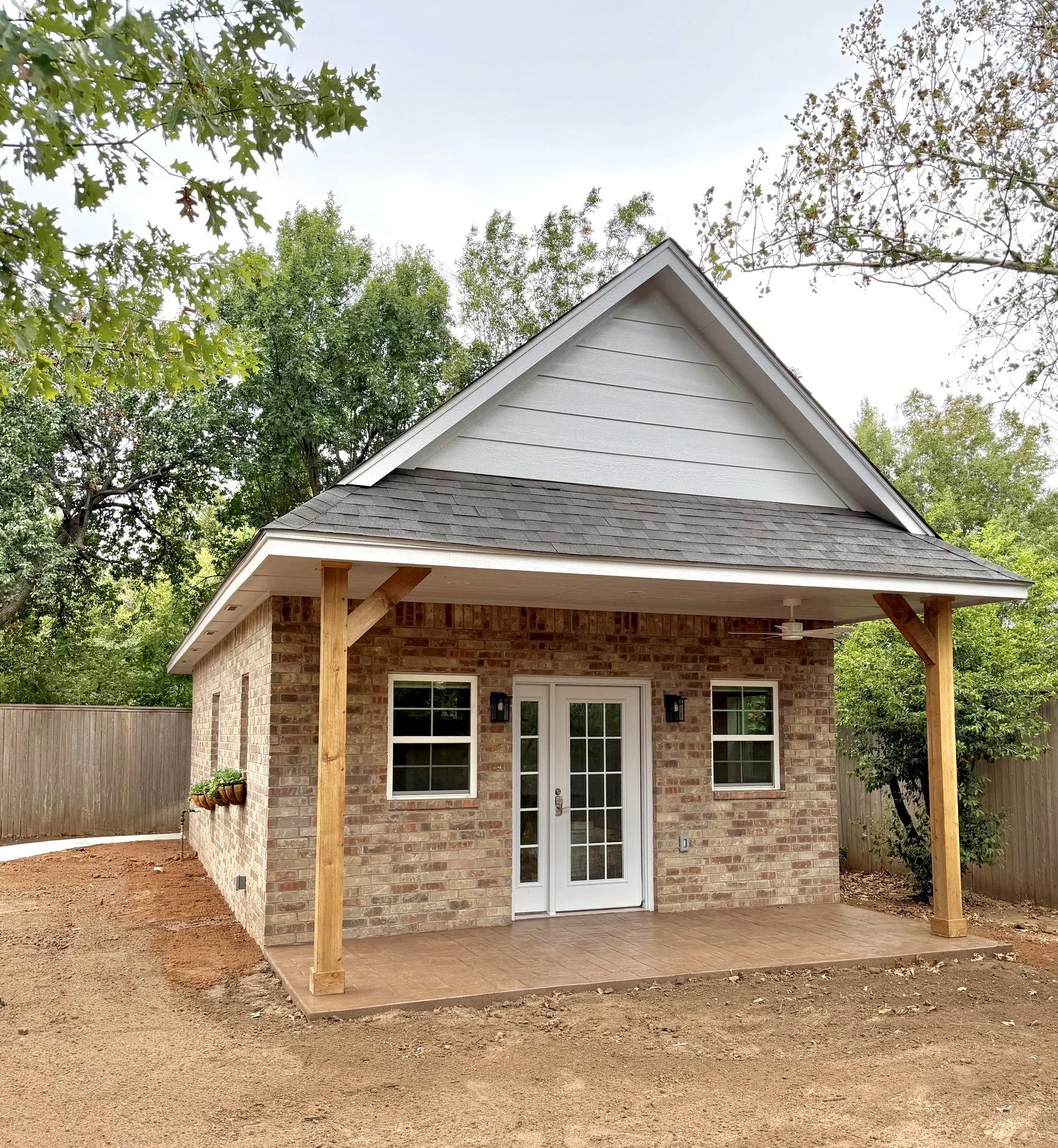 Backyard brick house with a newly built covered patio supported by wooden posts, white double door with glass panes, and two windows on either side. Surrounding trees and a partially landscaped yard.