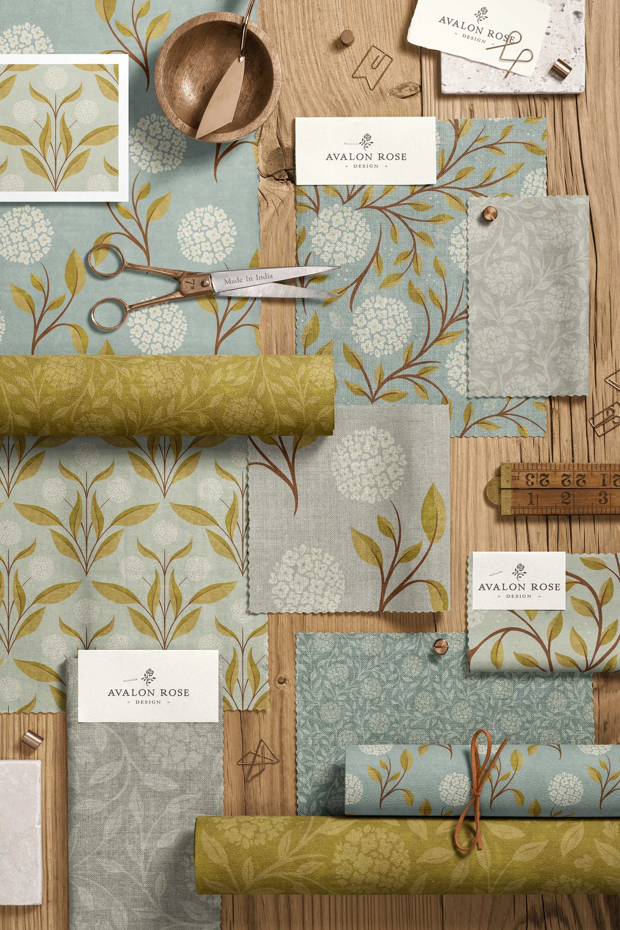 Flat lay of fabric swatches with floral patterns in shades of blue, green, and gray, gold-colored scissors, a wooden bowl, a measuring ruler, business cards with 'Avalon Rose Design,' and other decorative items on a wooden surface.