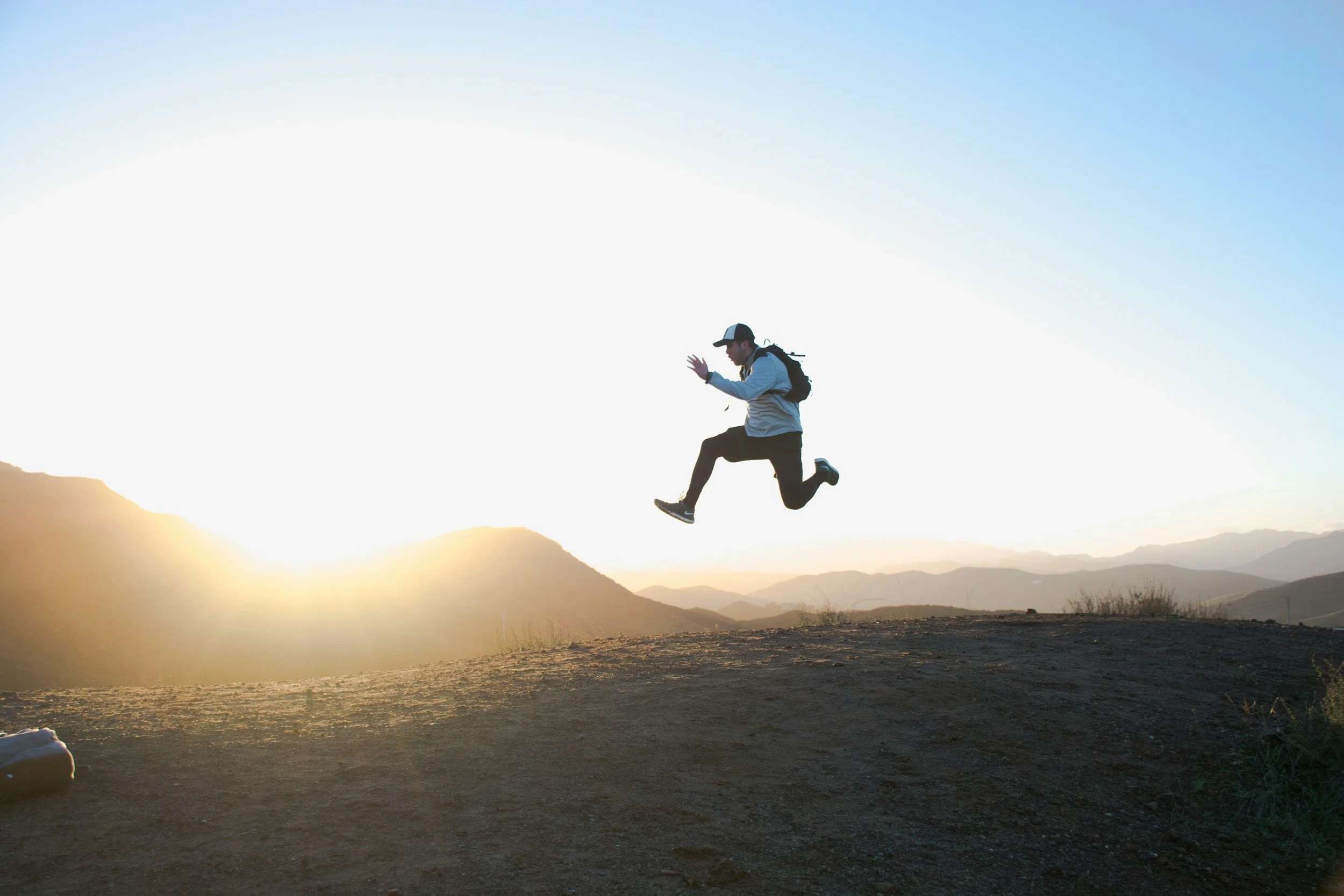Person jumping in the air on a mountain trail during sunset, with mountains in the background.
