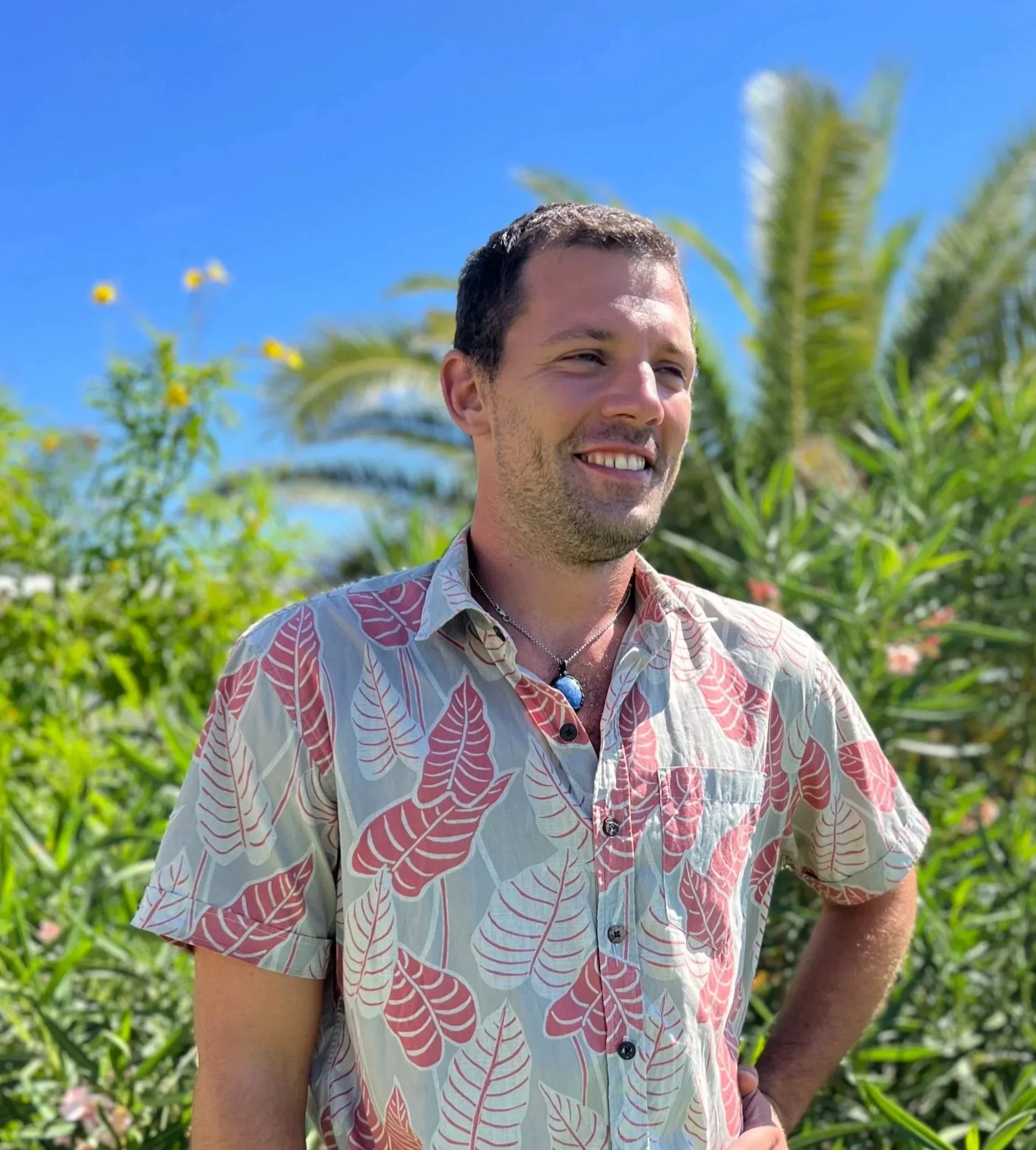 A smiling man with short dark hair, wearing a beige shirt with red and pink leaf patterns and a necklace with a blue pendant, standing outdoors among lush green plants and palm trees under a bright blue sky.
