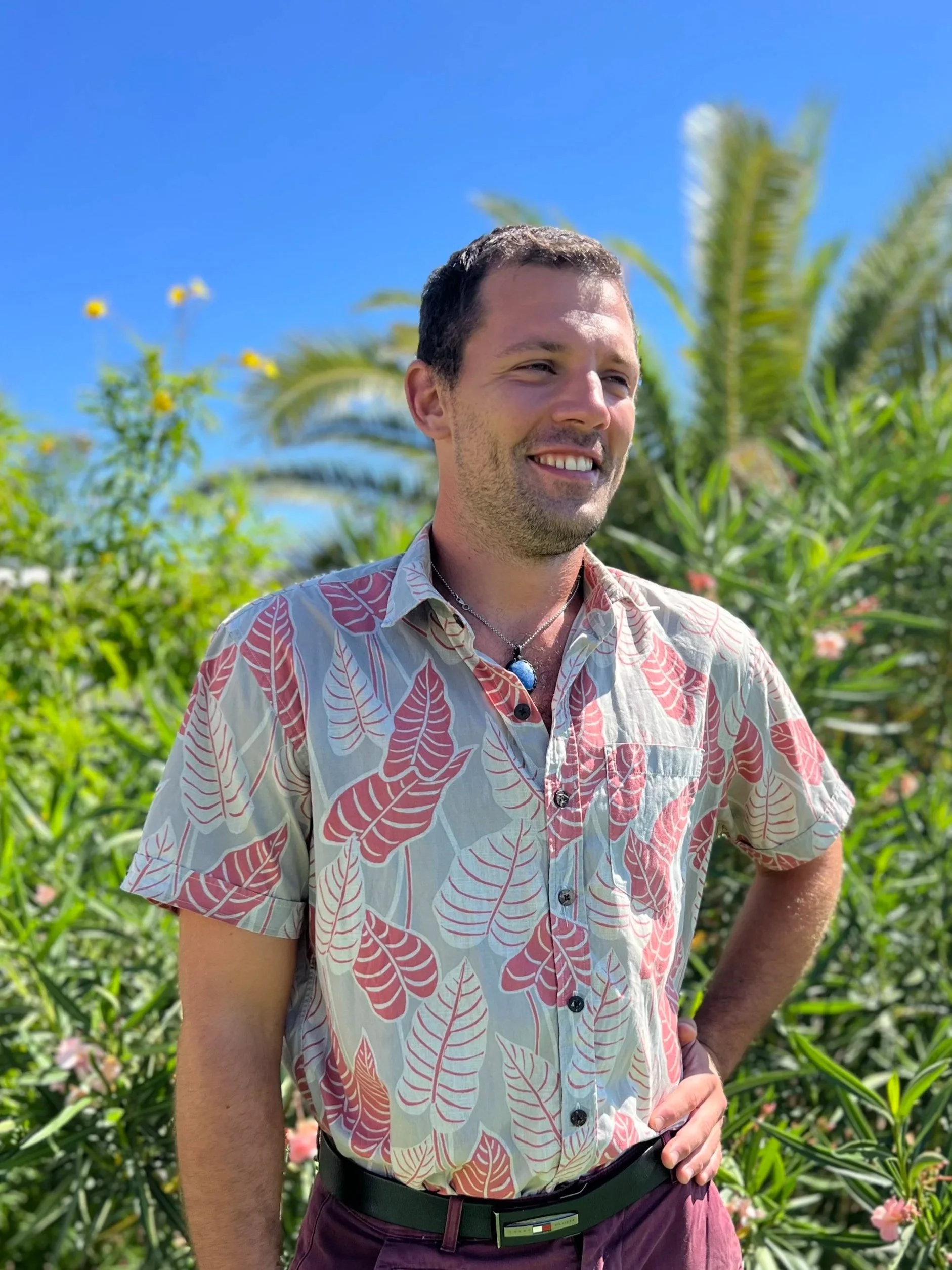 A man smiling outdoors in front of lush green plants and palm trees under a clear blue sky.