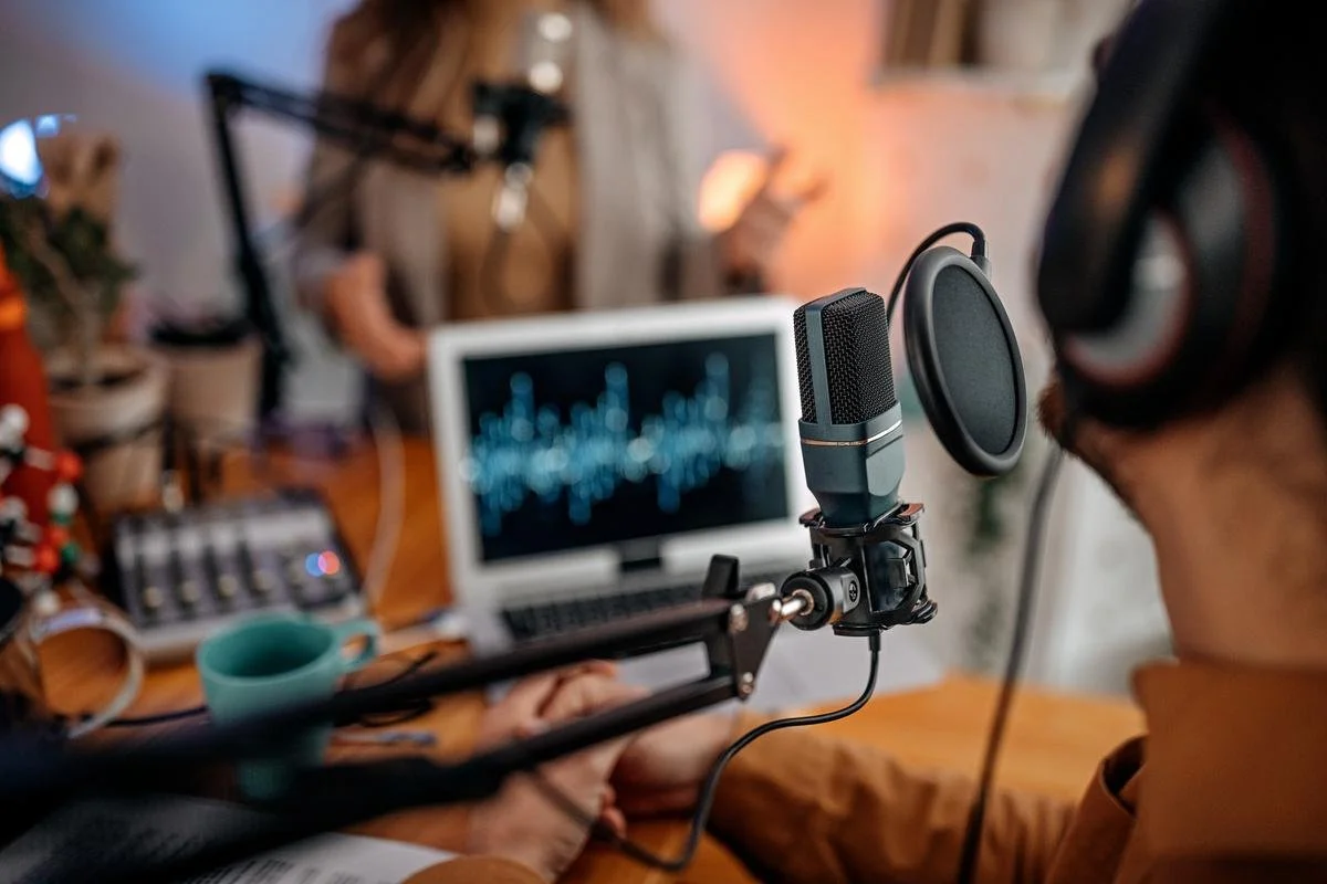 A person wearing headphones recording into a microphone in a home studio with a computer screen displaying audio waveforms in the background.