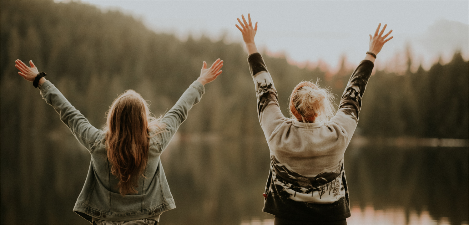 Two women stand by a lake with their arms raised in the air, facing away from the camera.