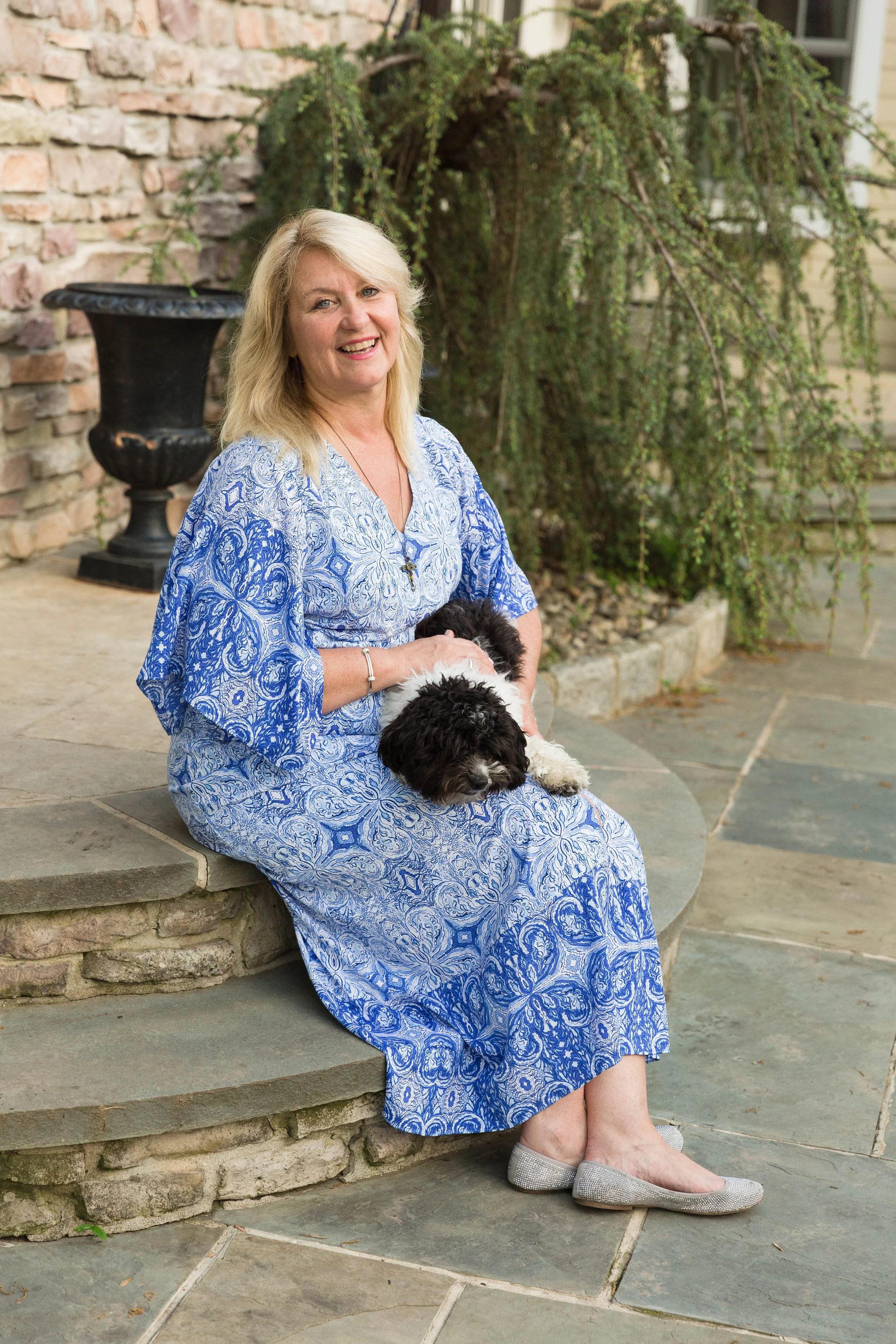 A woman with blonde hair and a blue patterned dress sitting on stone steps, holding a black and white dog, smiling outdoors with a stone wall, plants, and a window in the background.