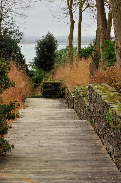 Jardin en pente à Cancale avec terrasse en bois, muret en pierre sèche, graminées aux couleurs d'automne et vue sur le littoral.