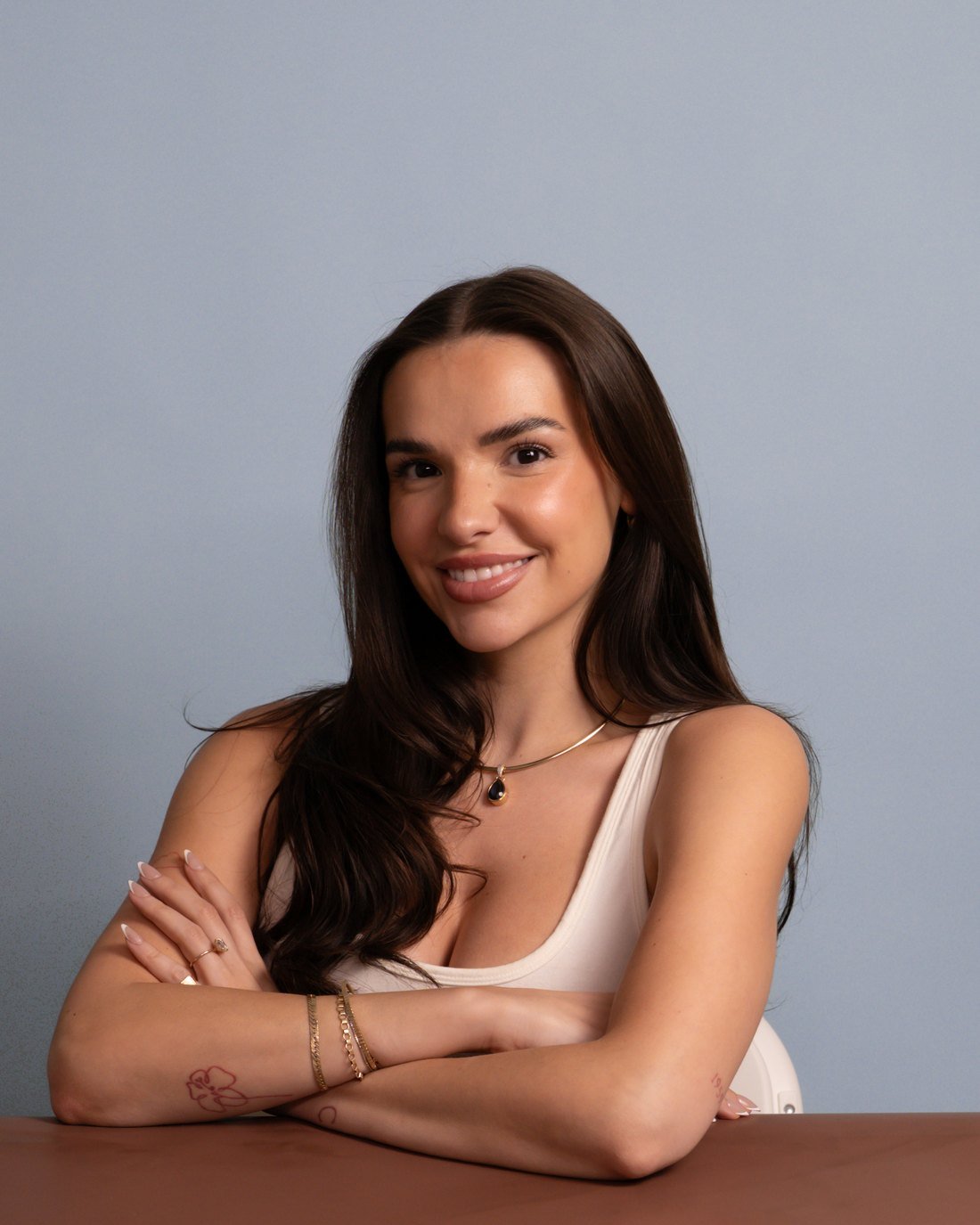 Portrait of Catherine Lockhart in a white tank with gold jewelry against a dusty blue backdrop