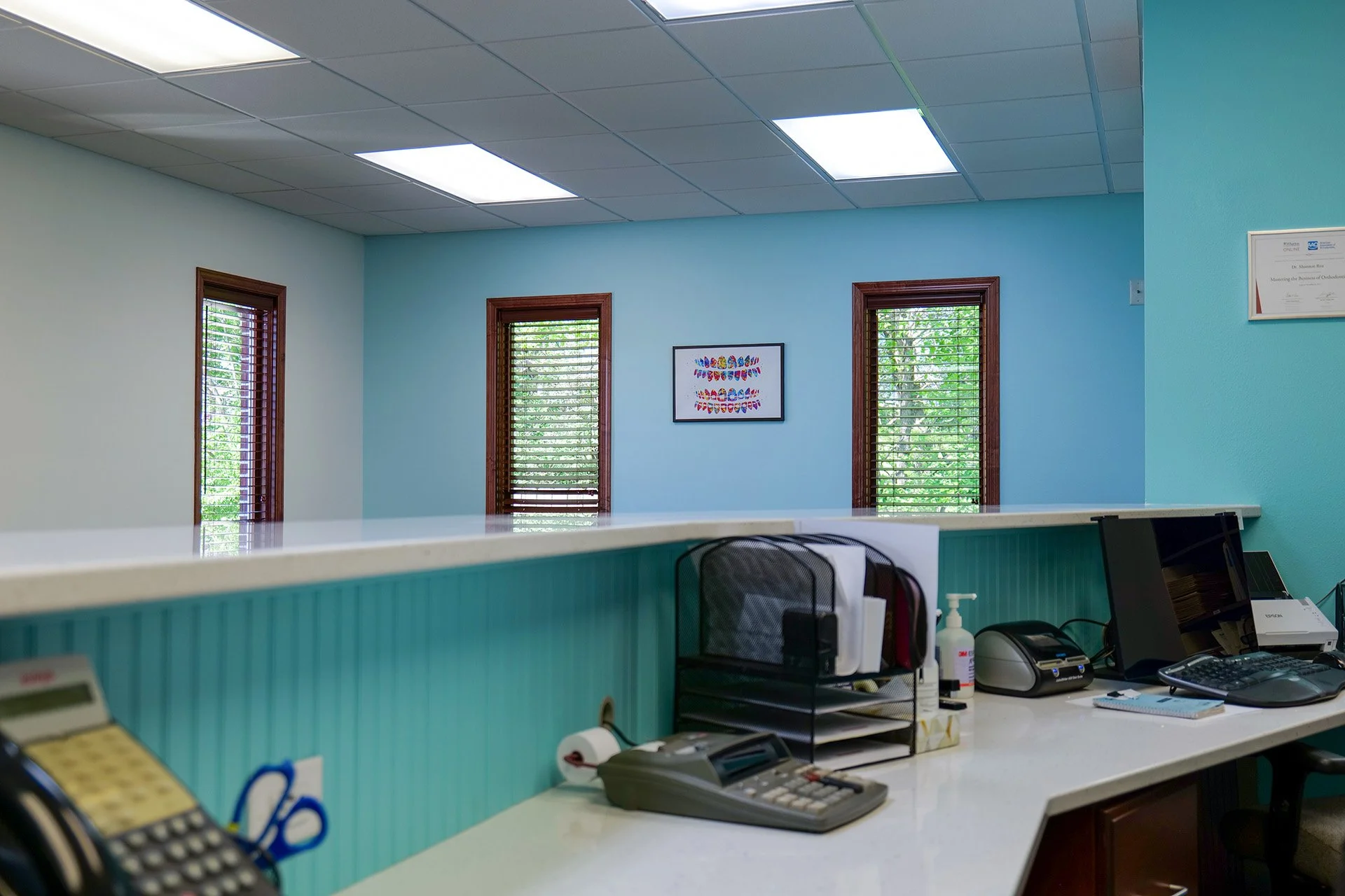 Office reception desk with teal paneling, computer equipment, and three windows with wooden frames showing green trees outside.