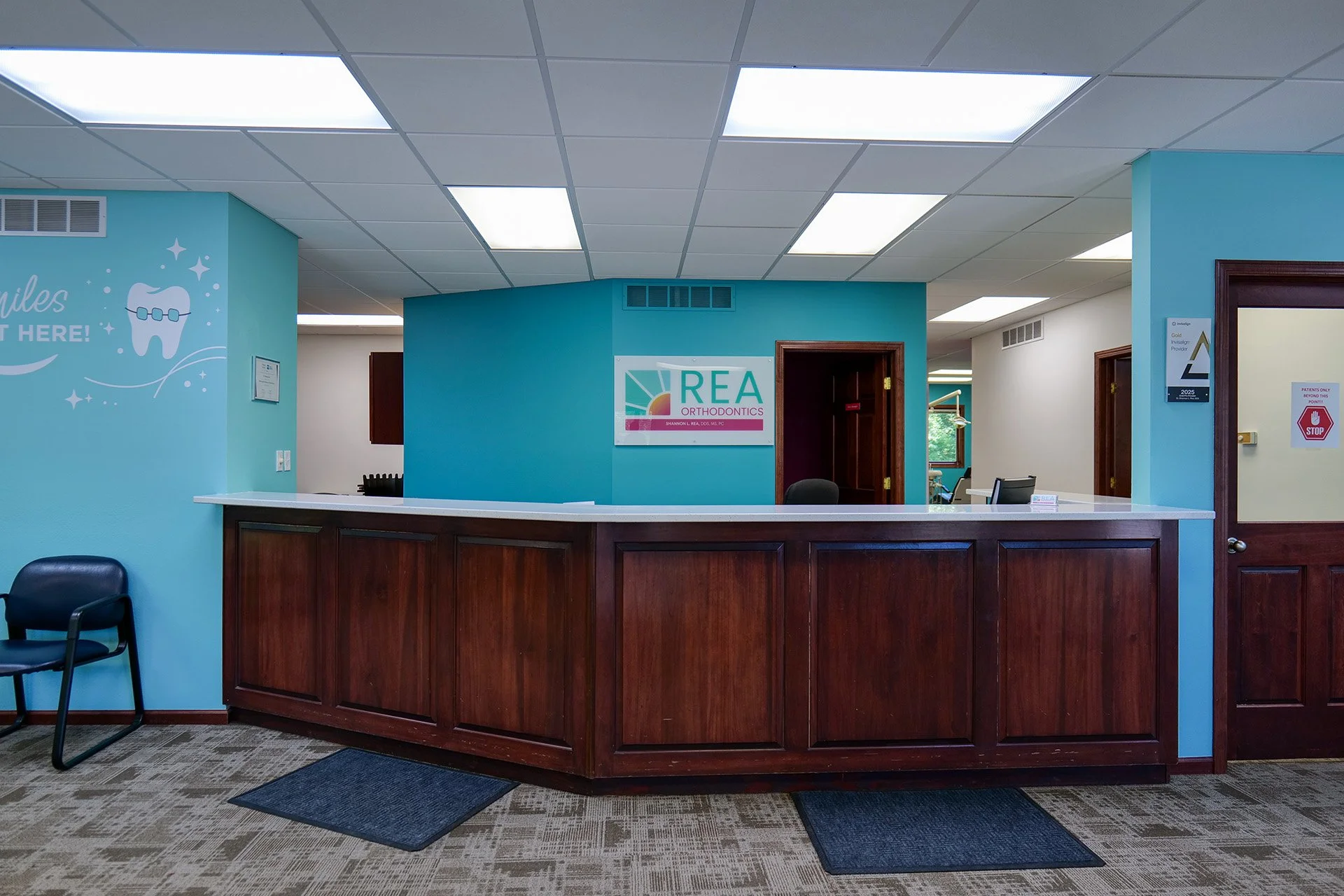 Reception desk at an orthodontics office with a turquoise wall, a sign reading "REA Orthodontics," and a cartoon tooth with a brace. There is a black chair and a doormat in front of the desk, and a partial view of a hallway with two doors.