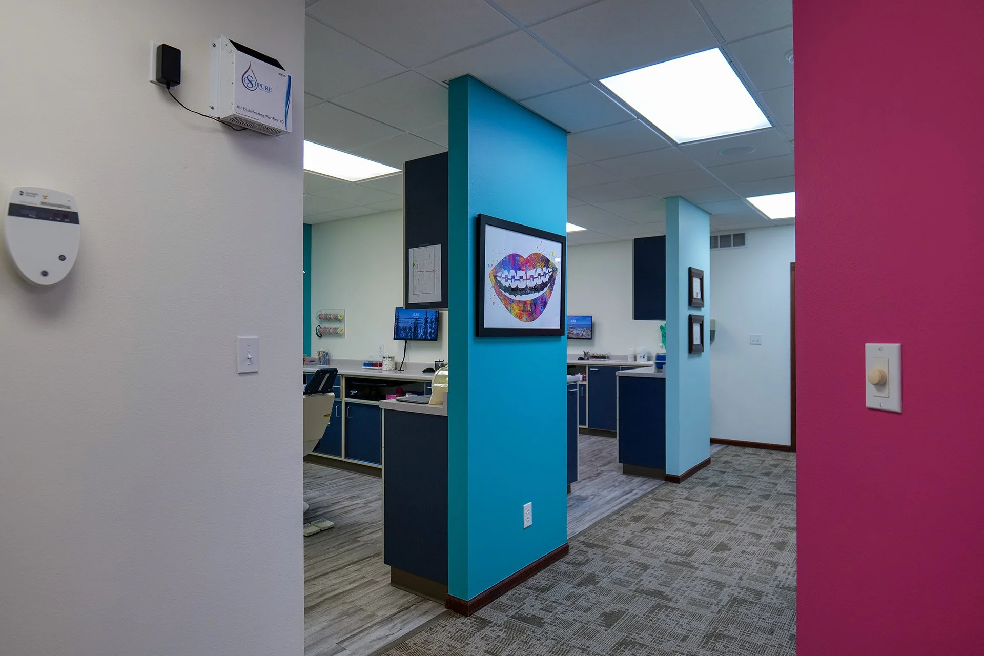 Interior view of a modern medical or dental office with colorful walls, including turquoise, navy blue, white, and pink. There are paintings, monitors, and office equipment visible.