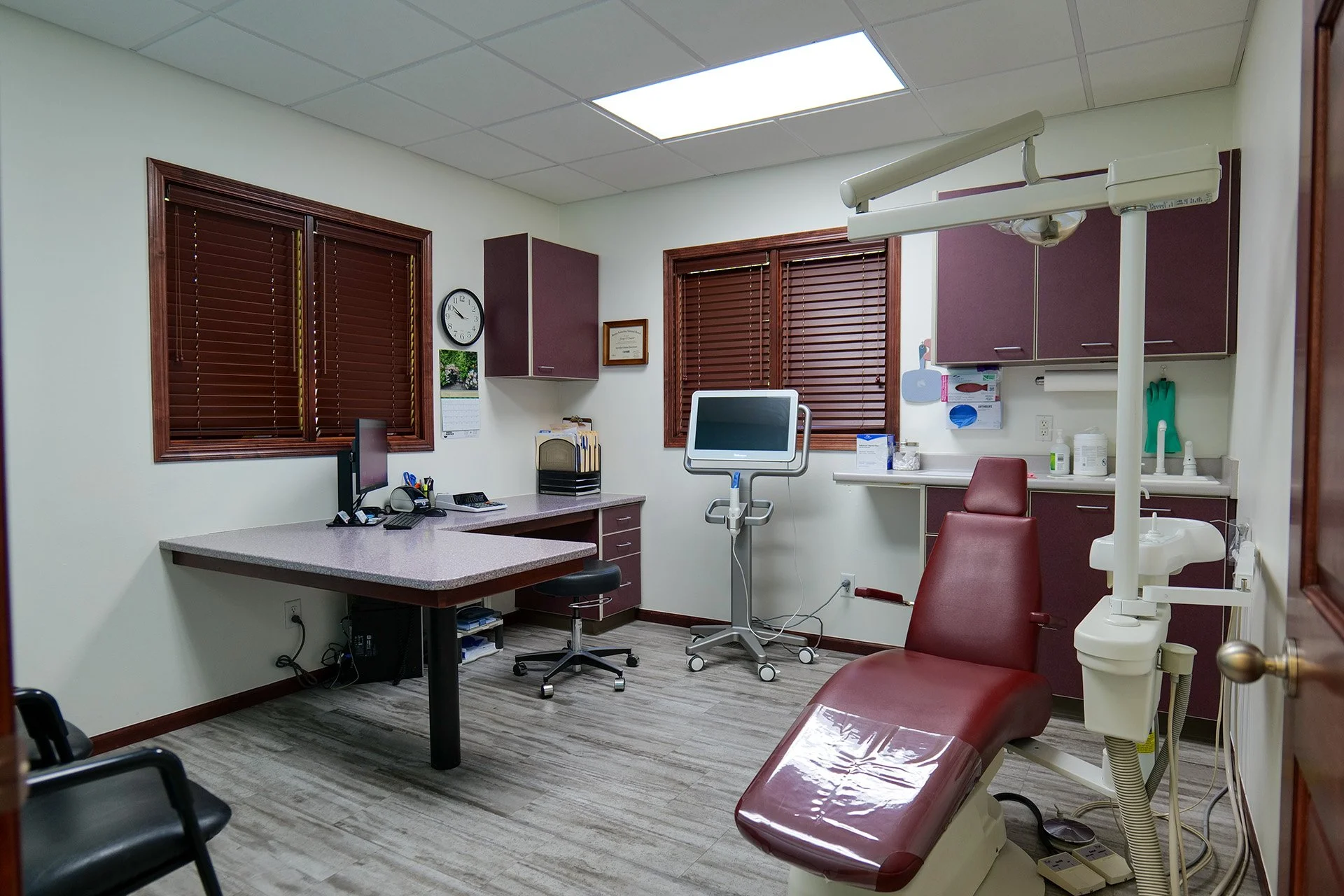 An empty dental examination room with wooden blinds on windows, a dental chair, a dental lamp, a computer, and medical supplies on countertops.