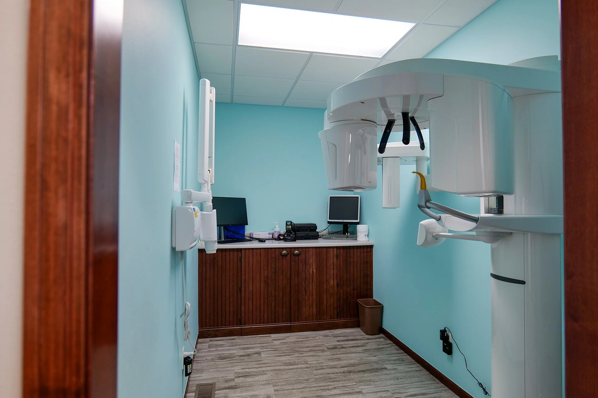 A dental X-ray machine in a dentist's office with light blue walls, a wood cabinet, a computer monitor, and various dental tools.