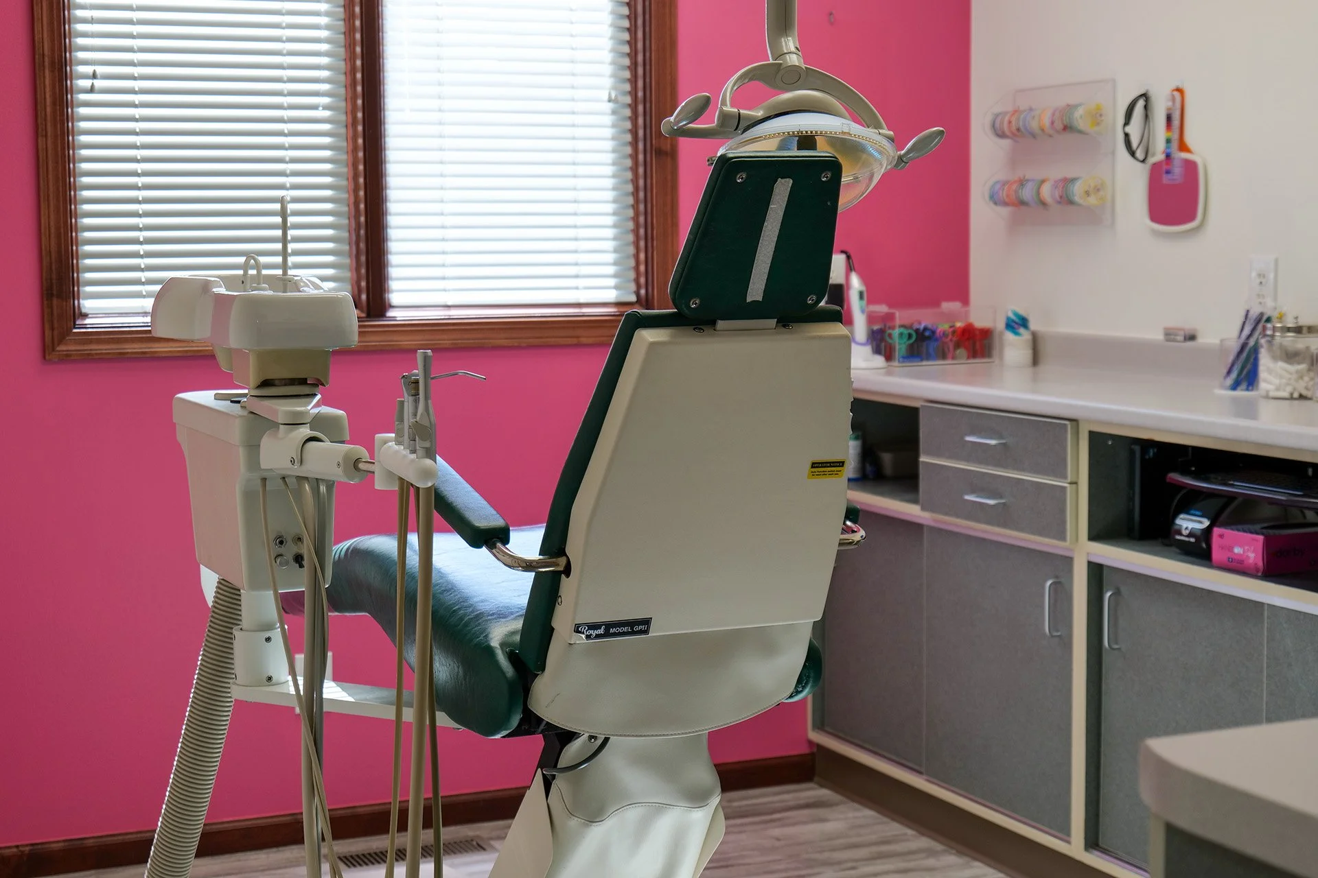 Dental examination room with dental chair, dental equipment, and supplies on counter and wall, pink wall, window with blinds.