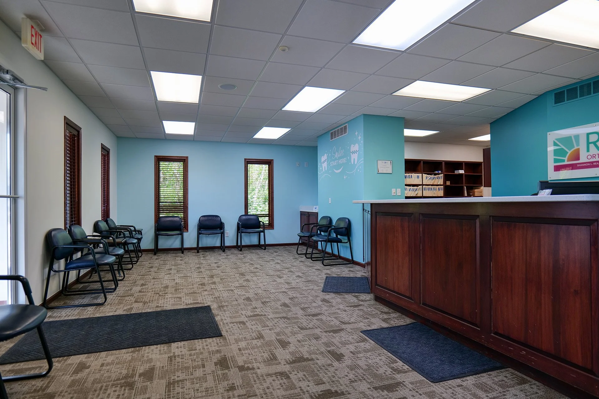 Waiting room area with black chairs along the wall, blue walls with windows, a wooden reception desk, and a wall with a dental smile art poster.