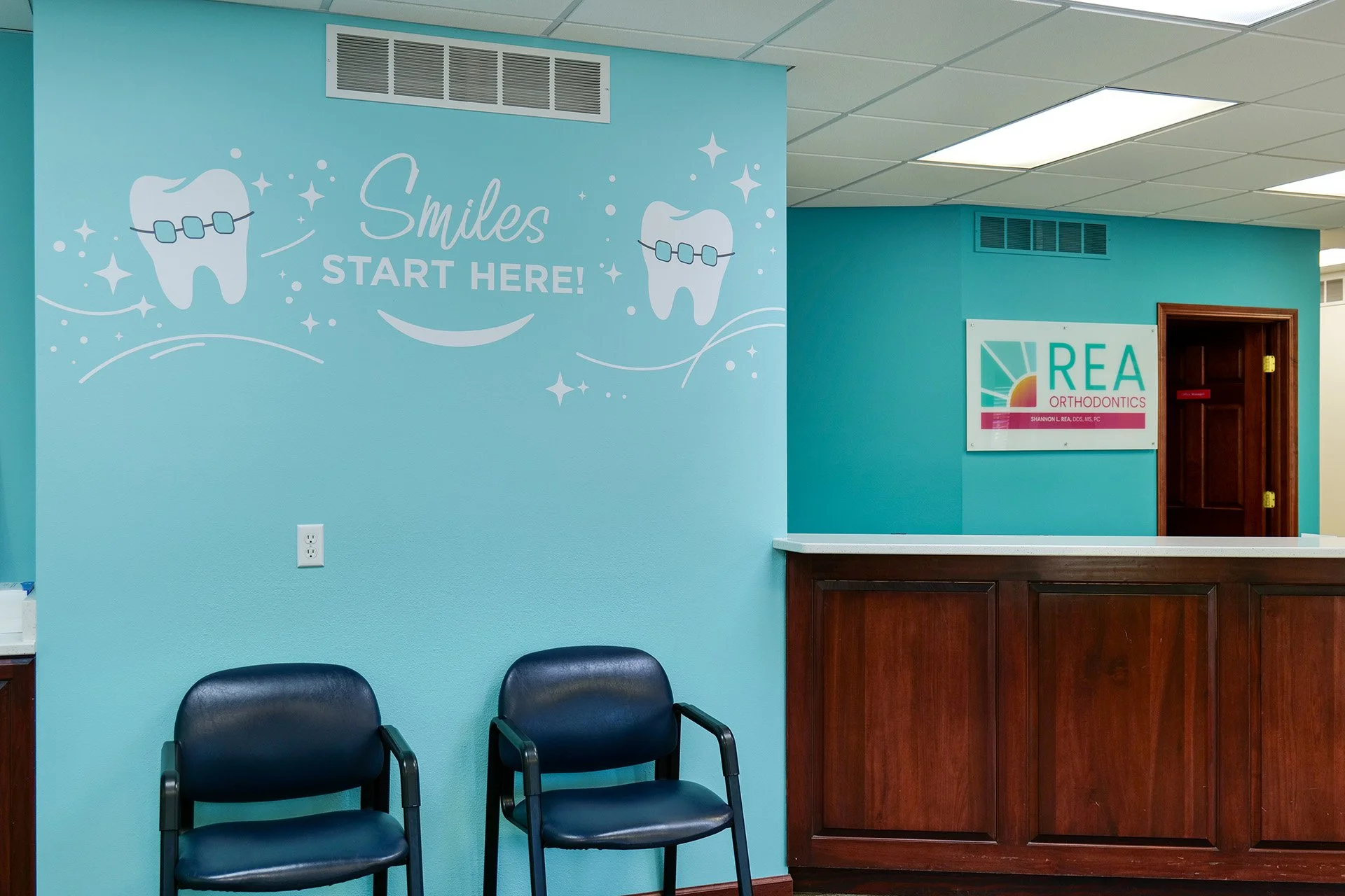 Waiting area with two black chairs and a light blue wall decorated with dental-themed graphics and the words "Smiles START HERE!" in a dental office reception area, near a tower with wooden panels and an orthodontics sign.