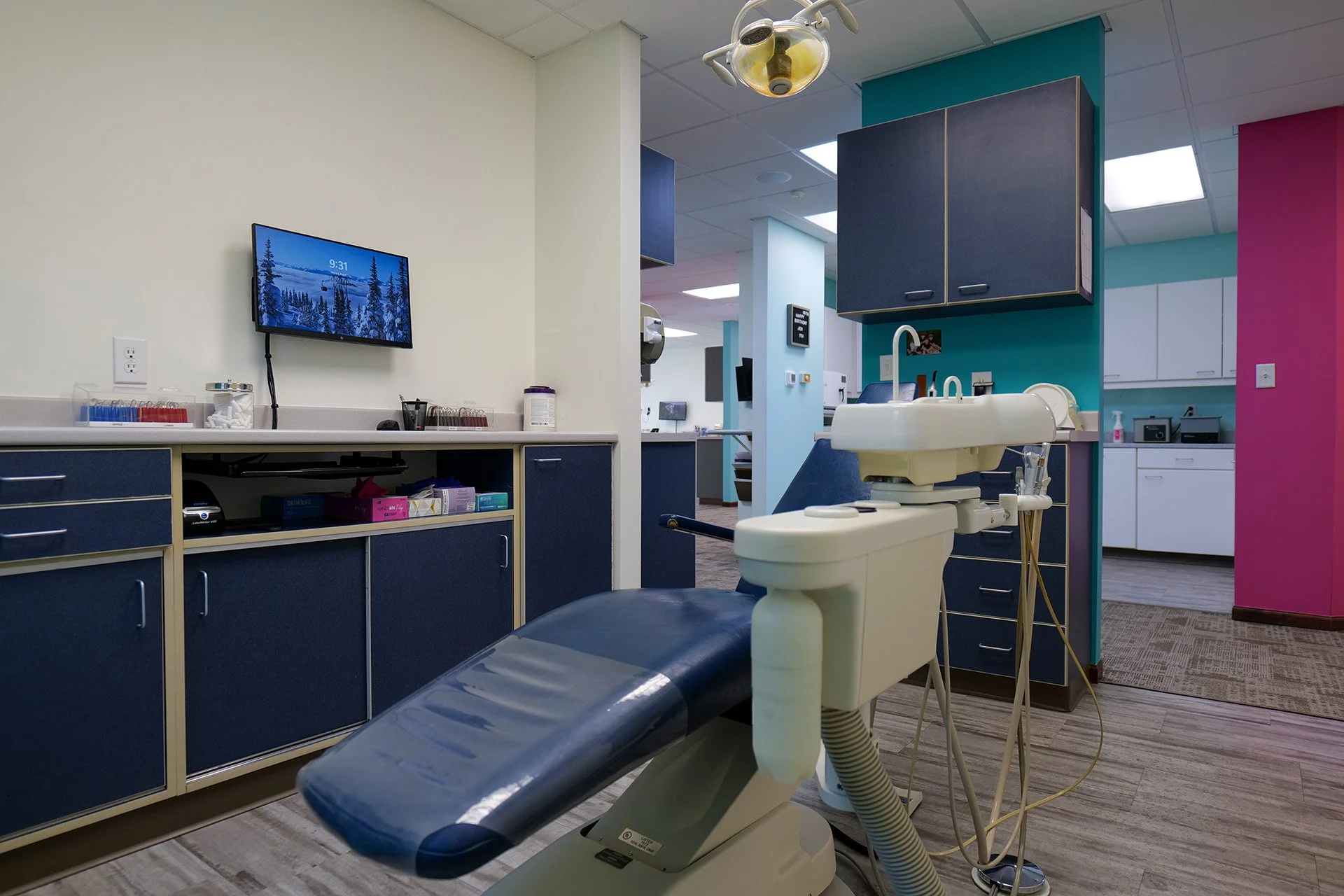 A dental office examination room with a dental chair, a white sink, and blue and pink walls. There are cabinets, a monitor on the wall displaying a winter landscape, and various dental supplies and equipment.