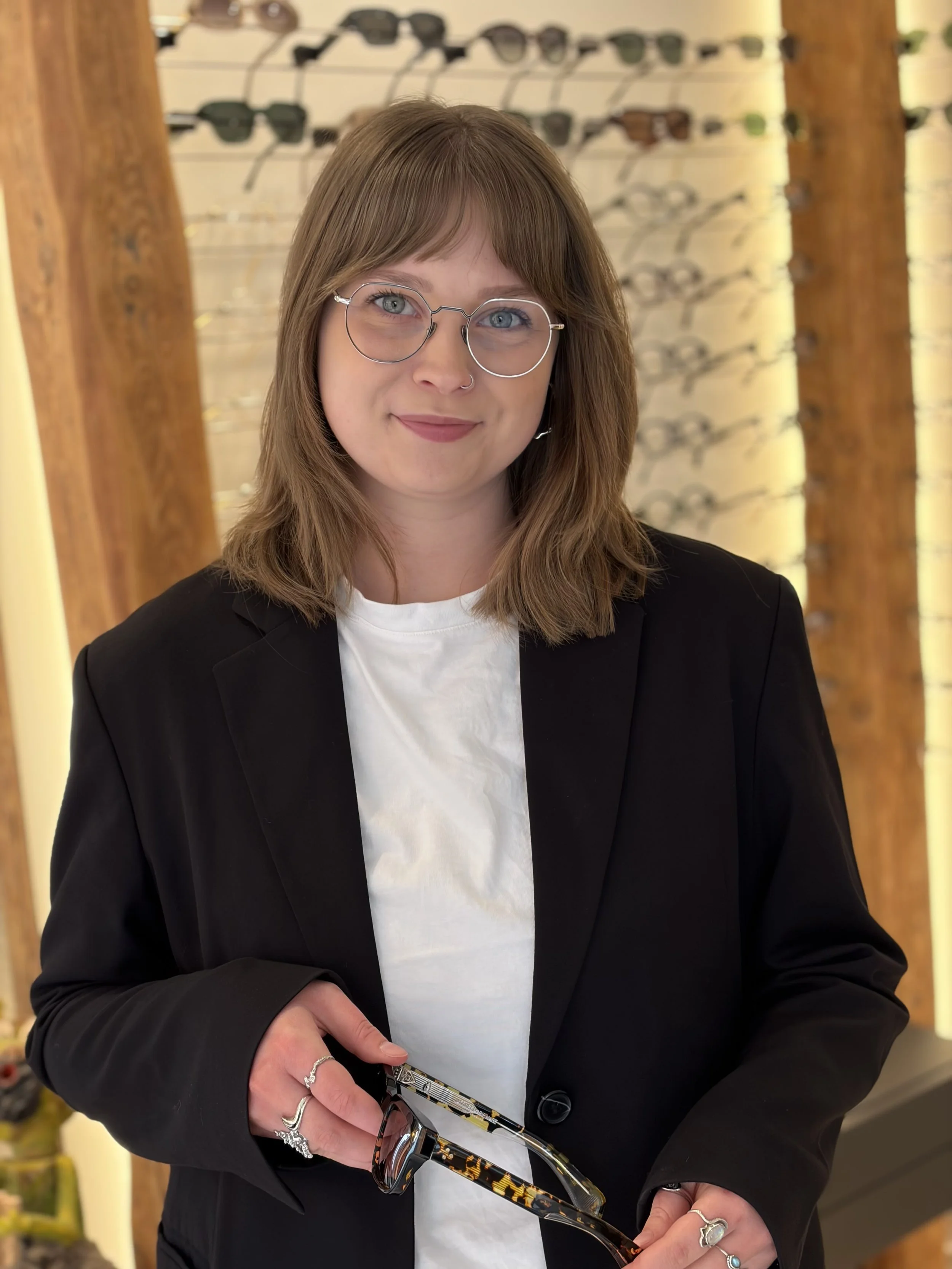Kvindelig person med brunt hår, iført brilller, sort blazer og hvid T-shirt, viser et par solbriller med tortoise print i en butik med solbriller på væggen bag hende.