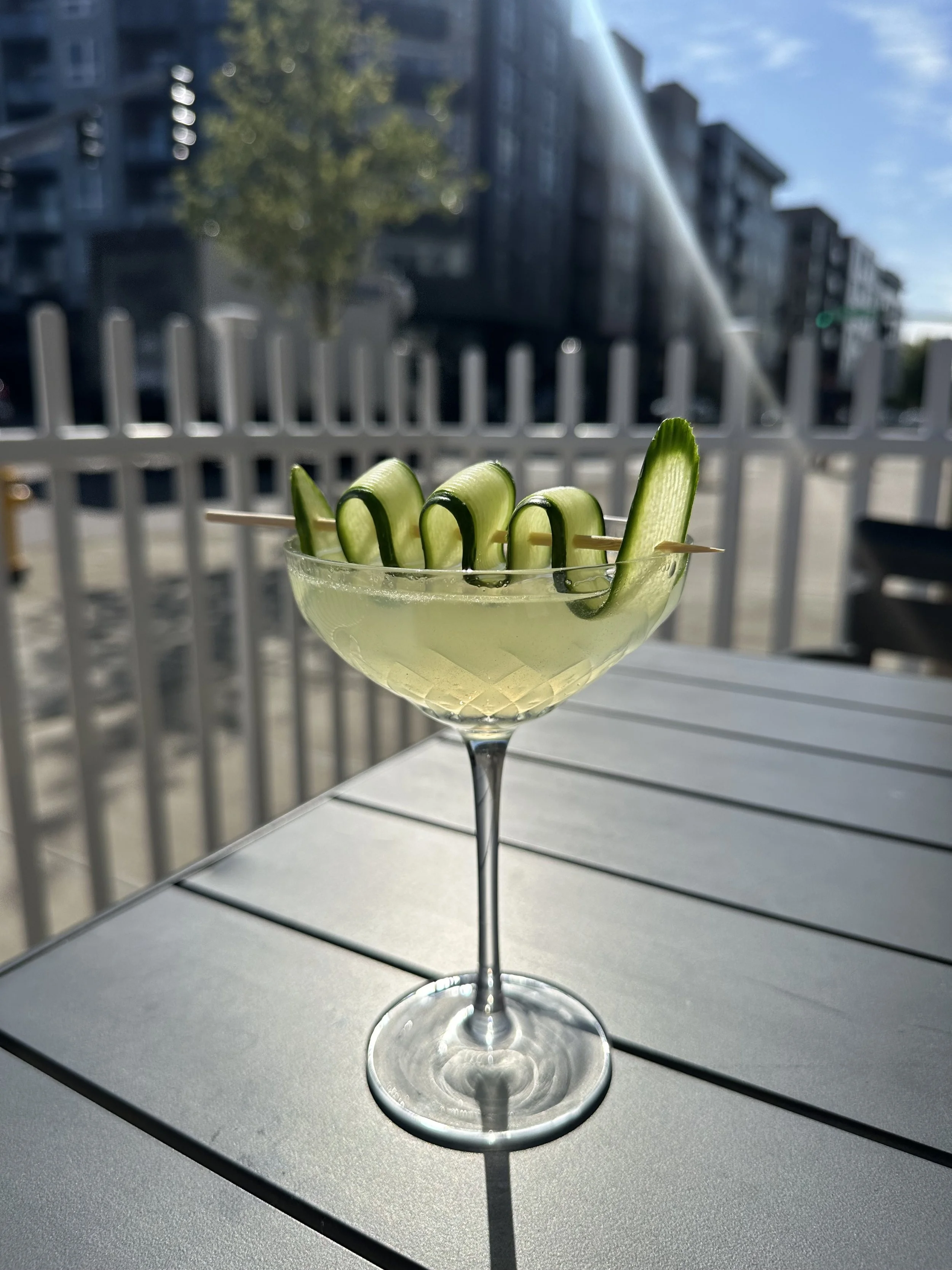 A cocktail glass with a cucumber garnish on the rim, placed on an outdoor table with a city street in the background.