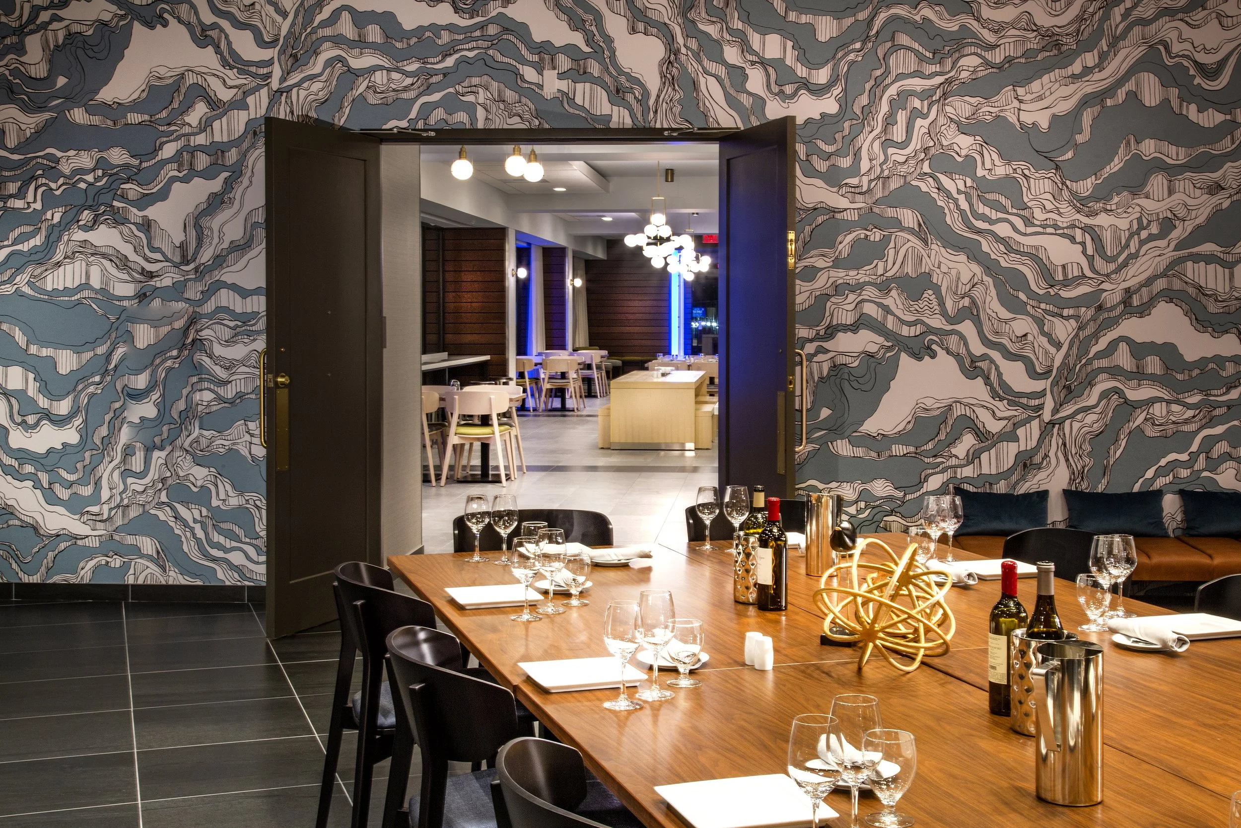 Elegant restaurant with a wooden table set with wine glasses, bottles, and decorative centerpiece, leading to a brightly lit dining area with modern lighting and wooden wall accents.