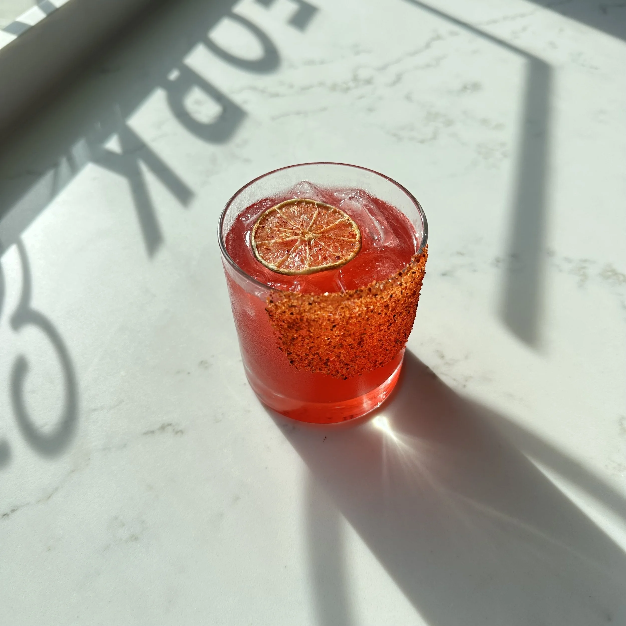 A pink cocktail in a clear glass with a slice of dried citrus fruit and a spiced rim, placed on a white marble surface with shadows from a window.