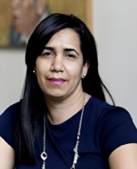 A woman with black hair and a dark blue shirt, smiling and wearing earrings and a necklace, in an indoor setting.