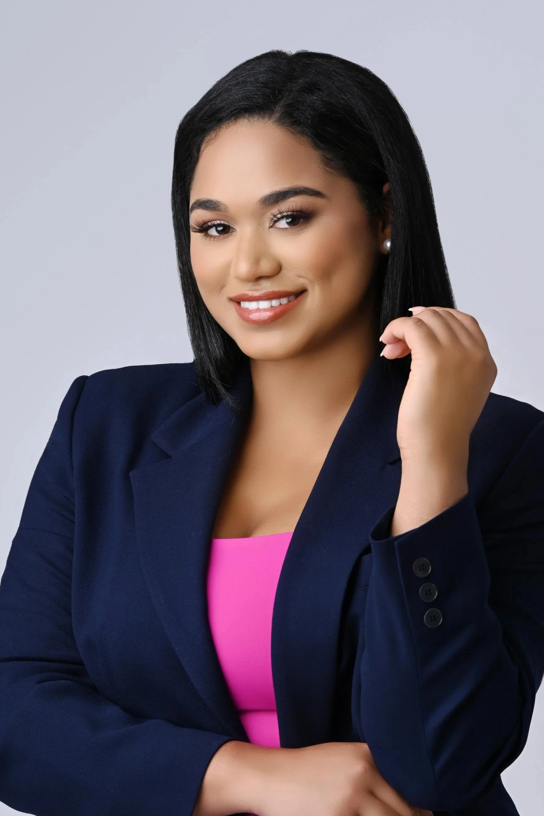 A woman with black hair wearing a navy blue blazer and a pink top, smiling at the camera against a plain gray background.