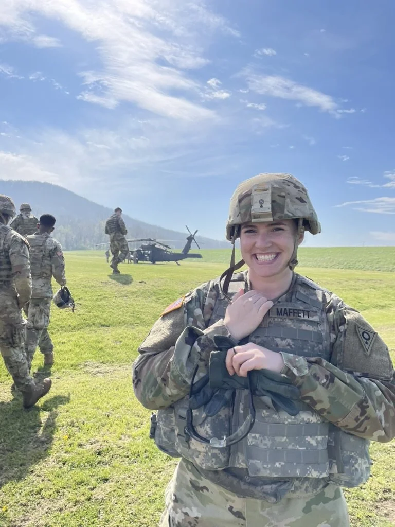 A smiling female soldier in camouflage uniform and helmet holding binoculars, with other soldiers and a helicopter in the background on a grassy field under a partly cloudy sky.