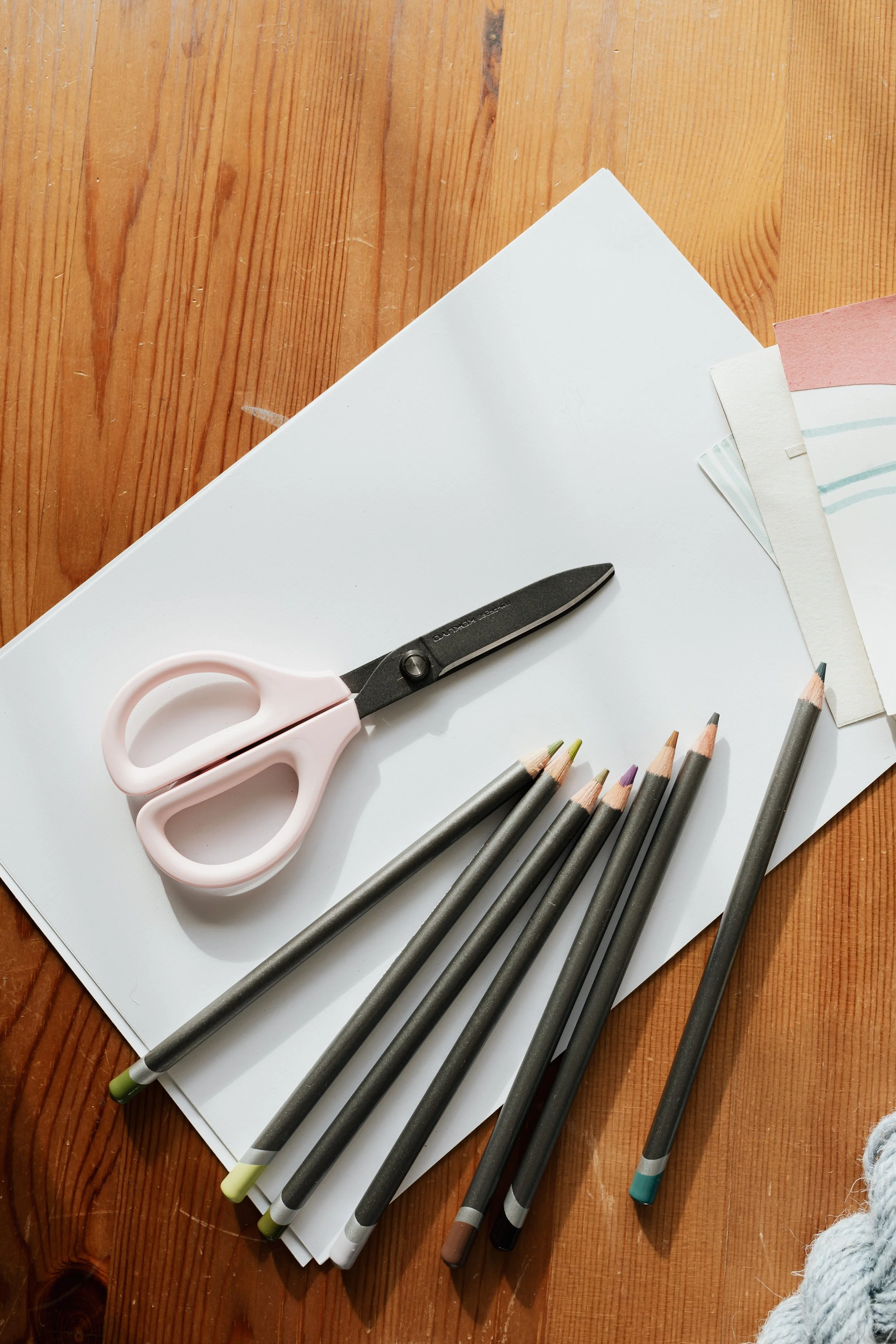 Une feuille de papier blanche posée sur une table en bois avec des crayons de couleur et des ciseaux roses, idéale pour créer un dopamine menu.
