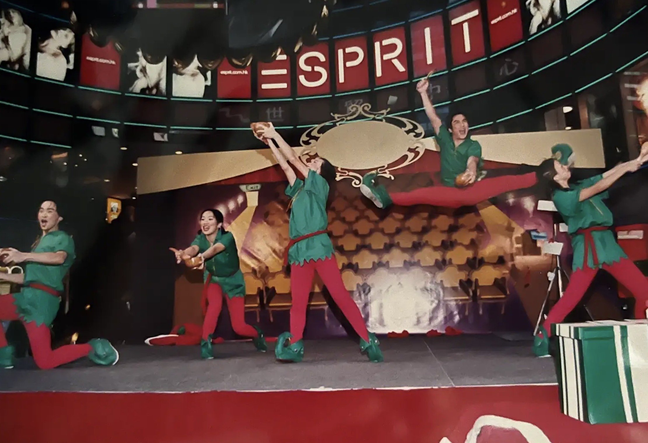 Group of five performers in festive costumes on stage, with some leaping and others holding objects, in front of a Christmas-themed background and oversized gift box, during a holiday show.