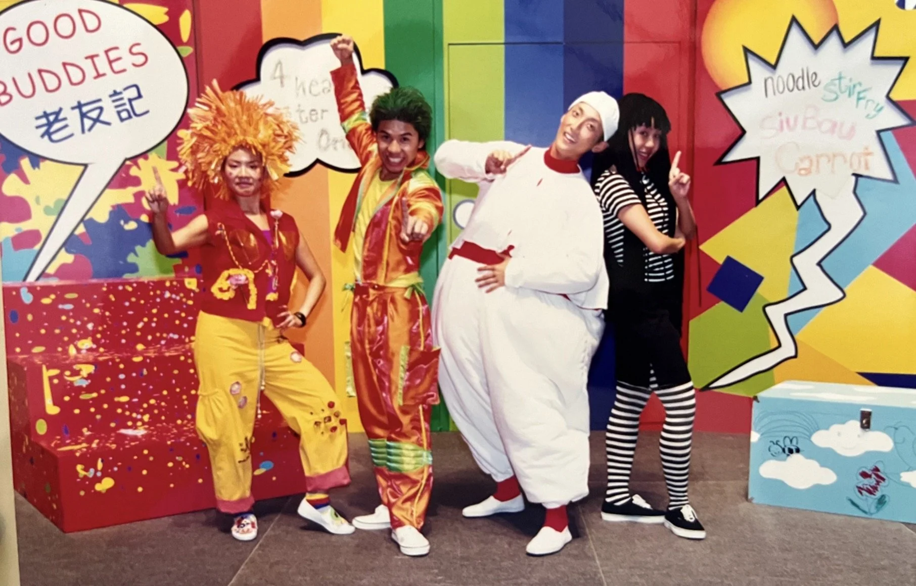 Four people in colorful costumes posing in front of a vibrant, comic-book style background with speech bubbles and graphics. The first person is dressed in a lion costume, the second in an orange and yellow outfit with green hair, the third in a chef's outfit with a white hat, and the fourth in black and white striped tights with black hair