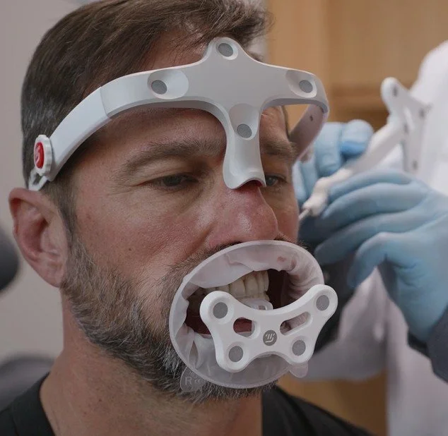 A man is having his mouth examined with a dental or medical device while wearing a headlamp and gloves, in a clinical setting.